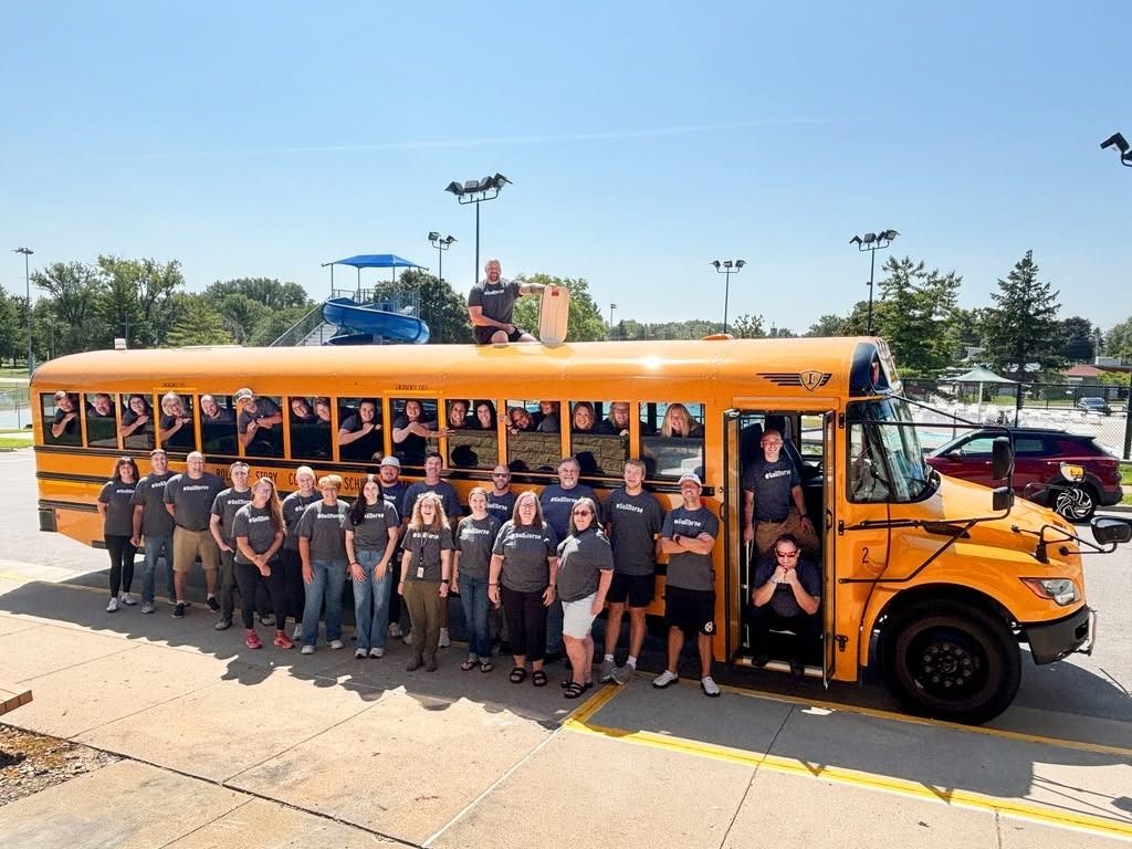 Group of people stand outside a yellow bus, some seated inside, on a sunny day.