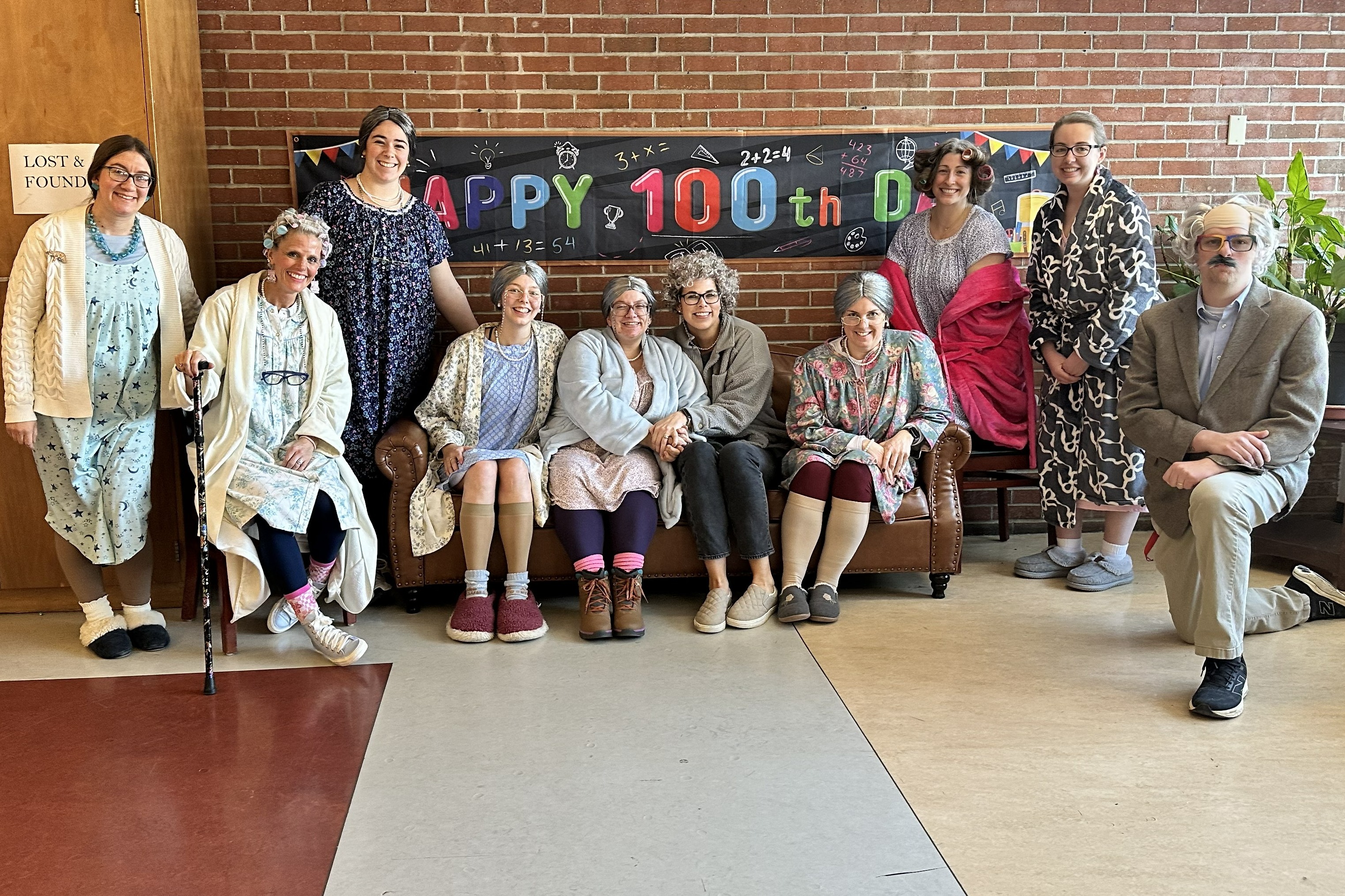 A group of school staff members dressed up as elderly individuals with gray wigs, robes, nightgowns, and walking canes pose together in front of a "Happy 100th Day" banner.