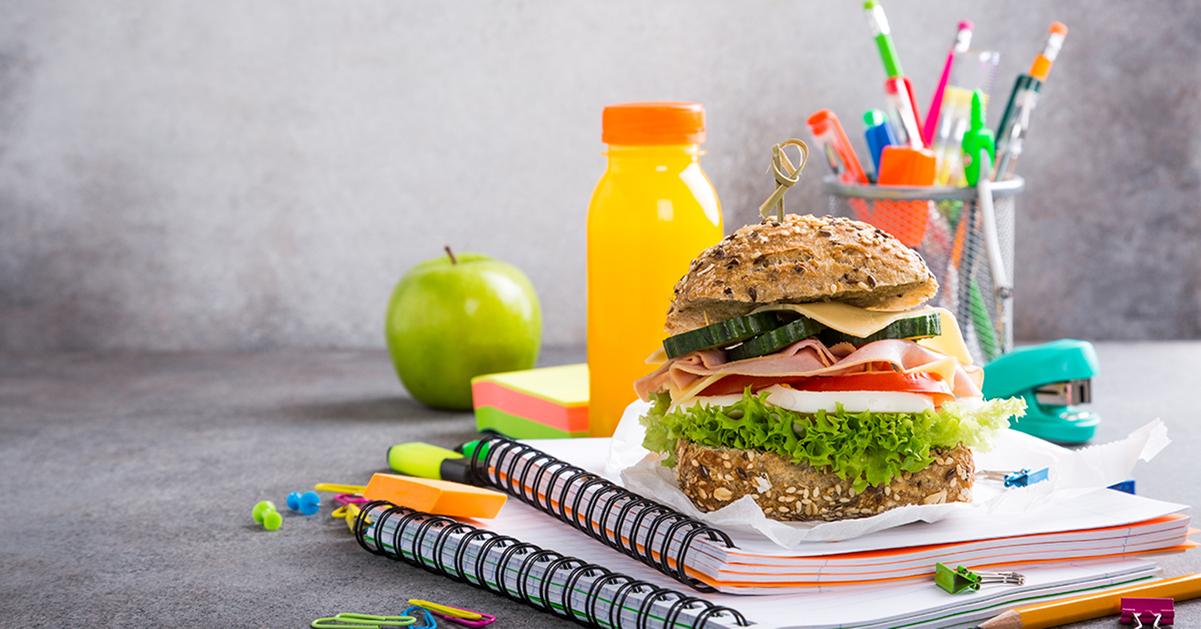 Sandwich on top of a notebook with an apple, orange juice, and pencils in background