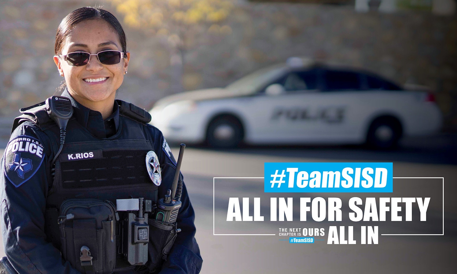 SISD Police Officer smiling with a police vehicle in the background, next to #TeamSISD All in for Safety
