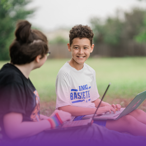 Two students sitting outside working on laptops and smiling at each other