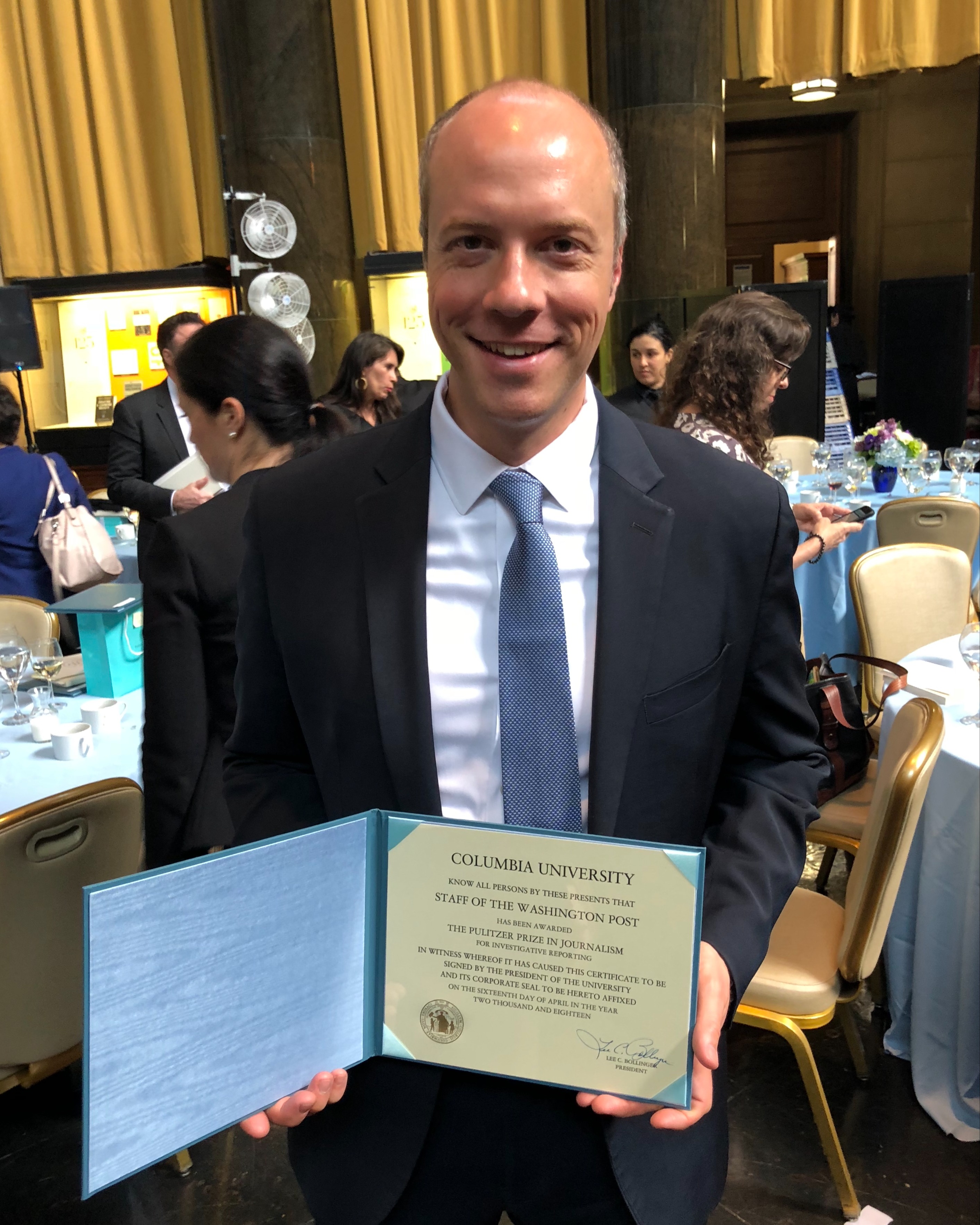 Picutre of alumi in a suite holding a diploma from Columbia