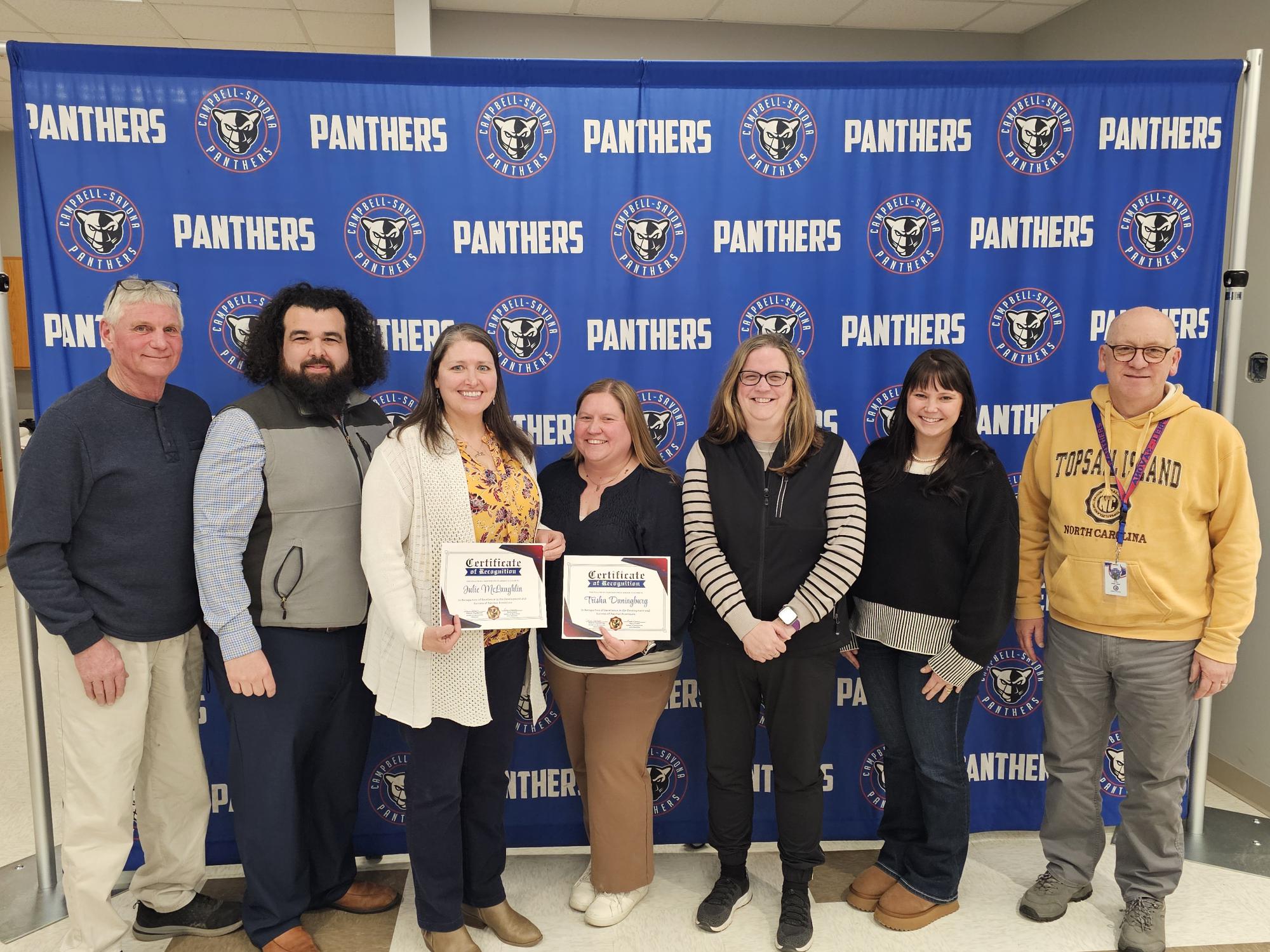 Board of education giving certificates of appreciation to two teachers standing in front of the panther banner