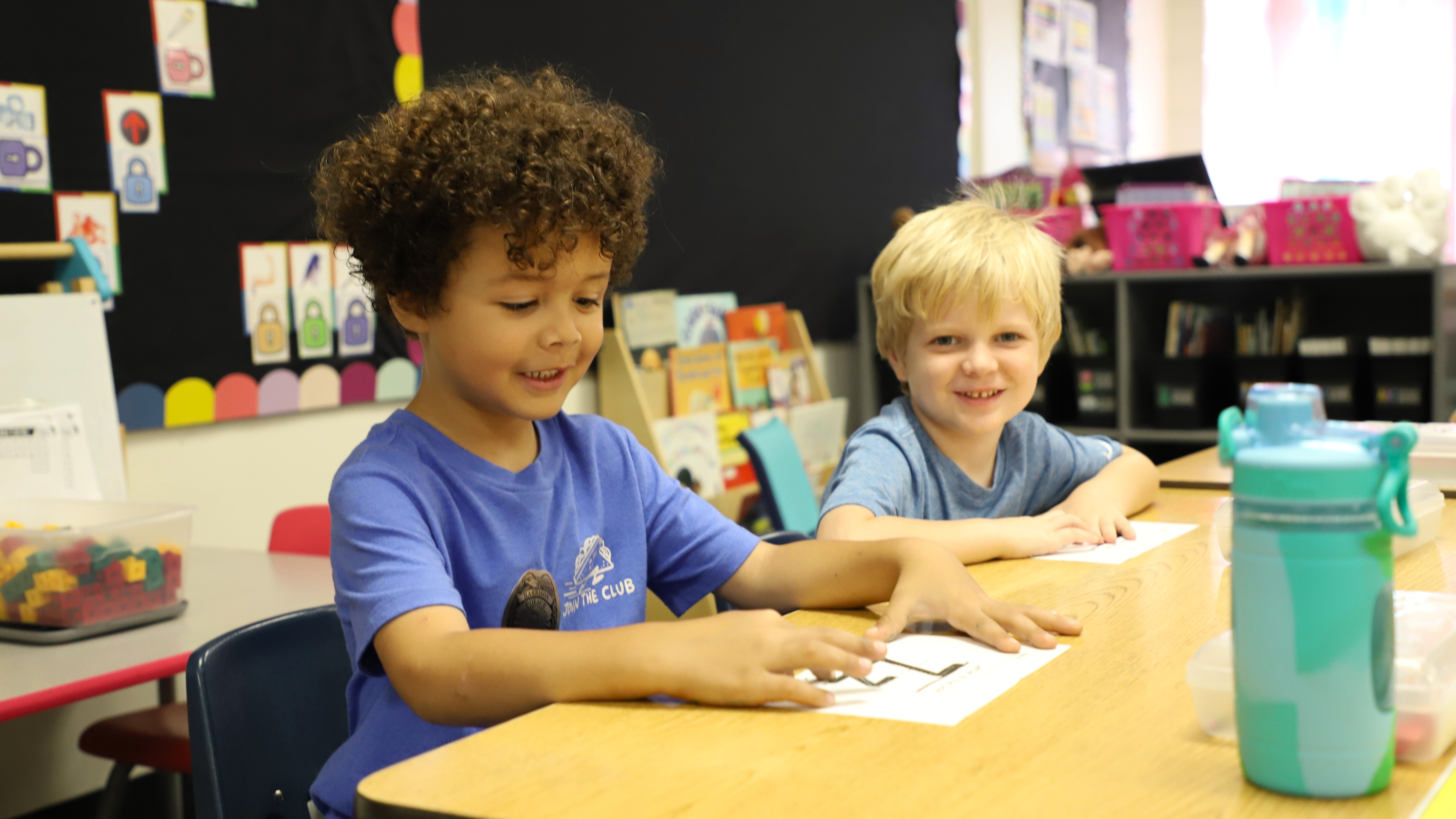 Two young boys sitting at a wooden desk in a classroom, with one working on a worksheet and the other smiling at the camera.