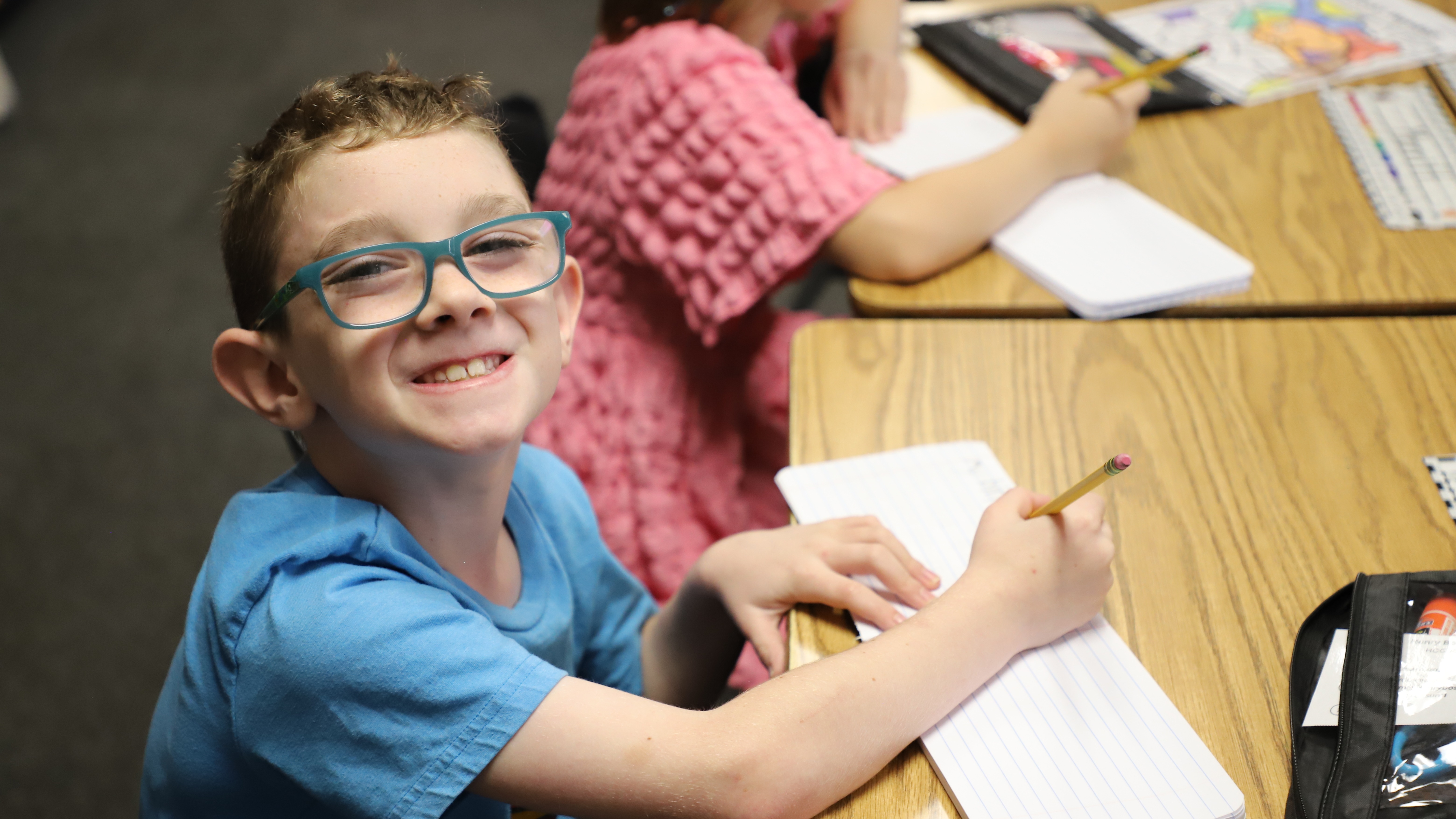 A young boy with glasses sitting at a classroom desk, smiling at the camera while holding a pencil over a notebook.