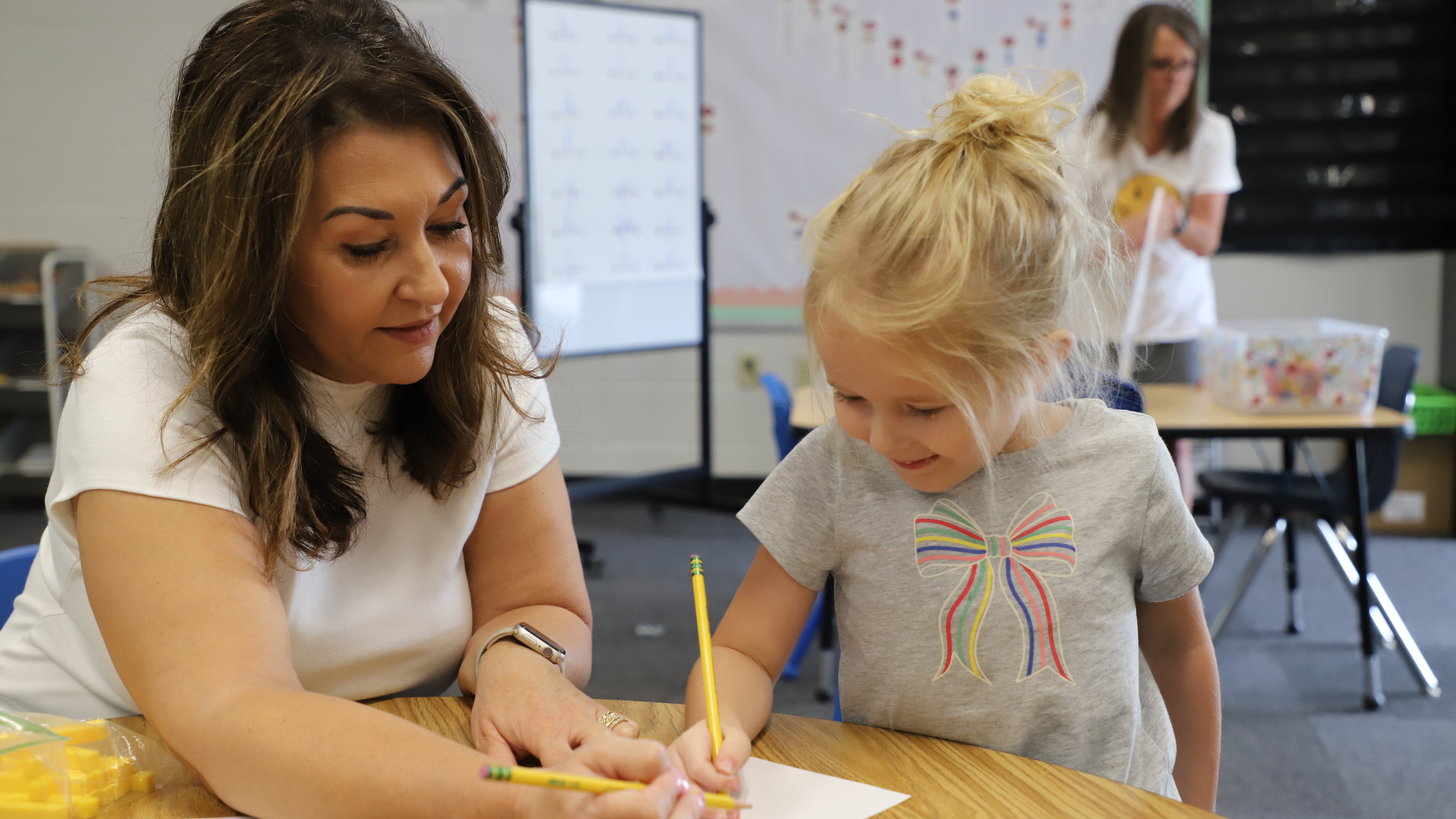 A teacher sits beside a young girl with blonde hair tied in a messy bun, guiding her as she writes on a paper with a pencil.
