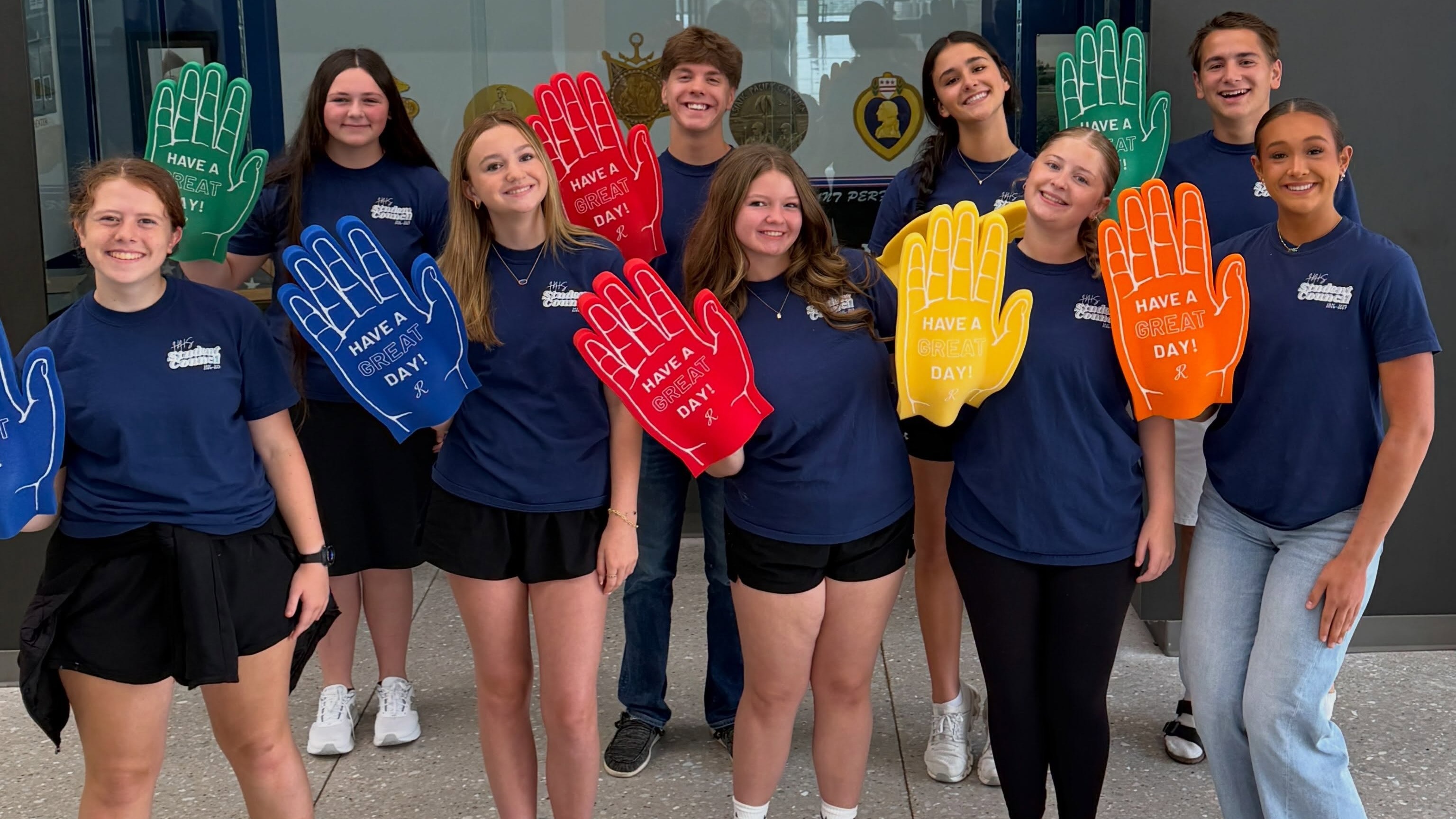 A group of nine smiling students wearing matching navy blue shirts standing in a hallway, holding up large foam high-five hands in various bright colors.