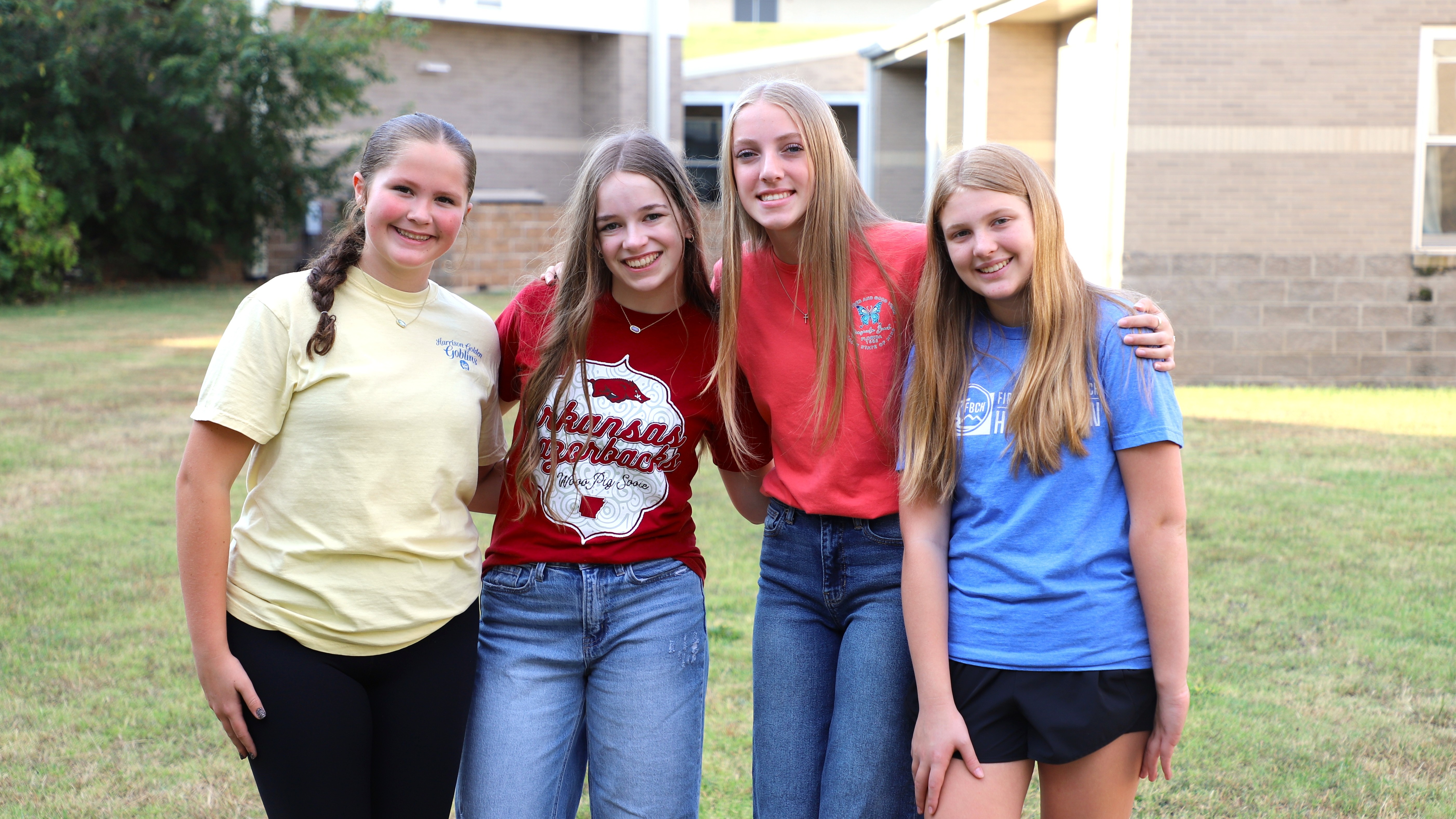 Four smiling teenage girls standing side-by-side on a grassy field in front of a school building, posing with their arms around one another.