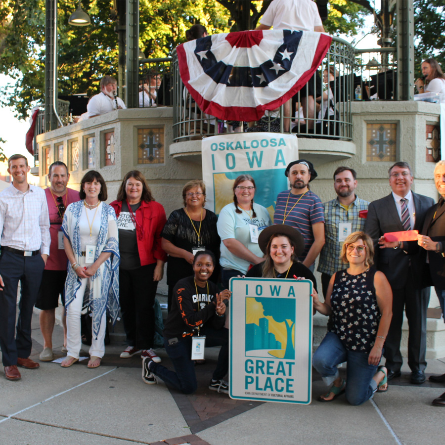 group of individuals gathered around an Iowa Great Place sign at the Oskaloosa Bandstand