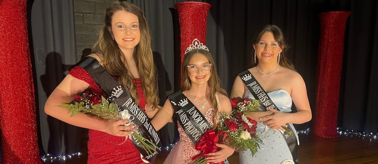 Three girls wearing pageant dresses pose on a stage.