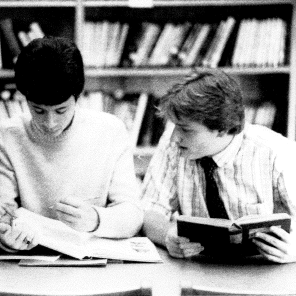 1985 Library. 3 people at a desk reading and talking