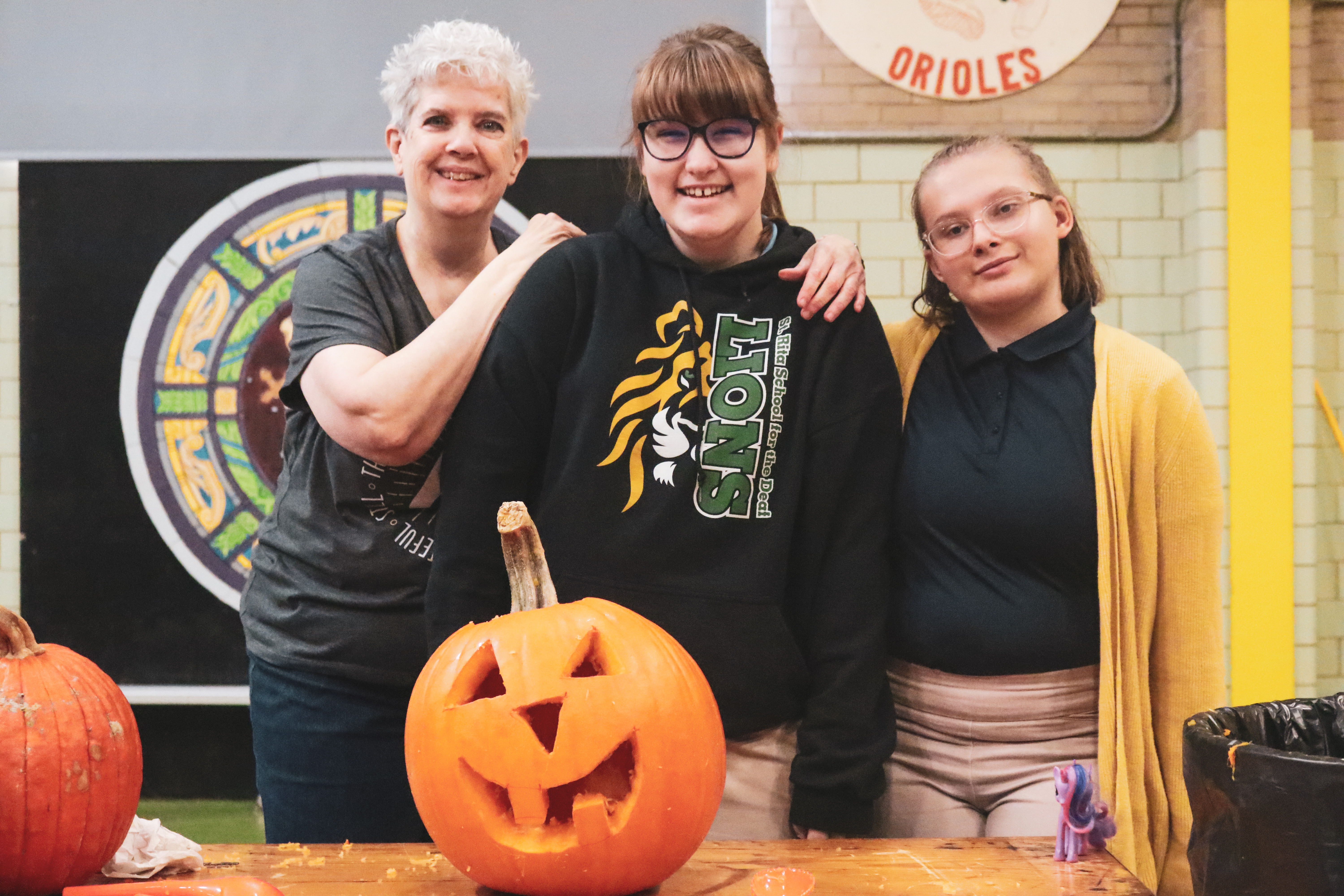 a teacher with two female students smiling with a carved pumpkin
