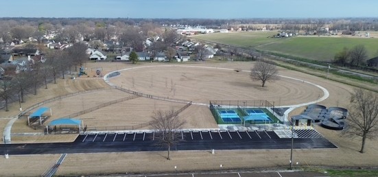 Aerial view of a large park under development, featuring a circular walking path, newly built pickleball courts, a pump track, parking area, playground equipment, and open grassy space surrounded by a neighborhood.