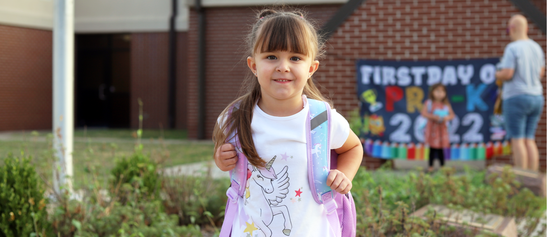 A preschool age girl in a unicorn shirt smiling at the camera on the first day of school