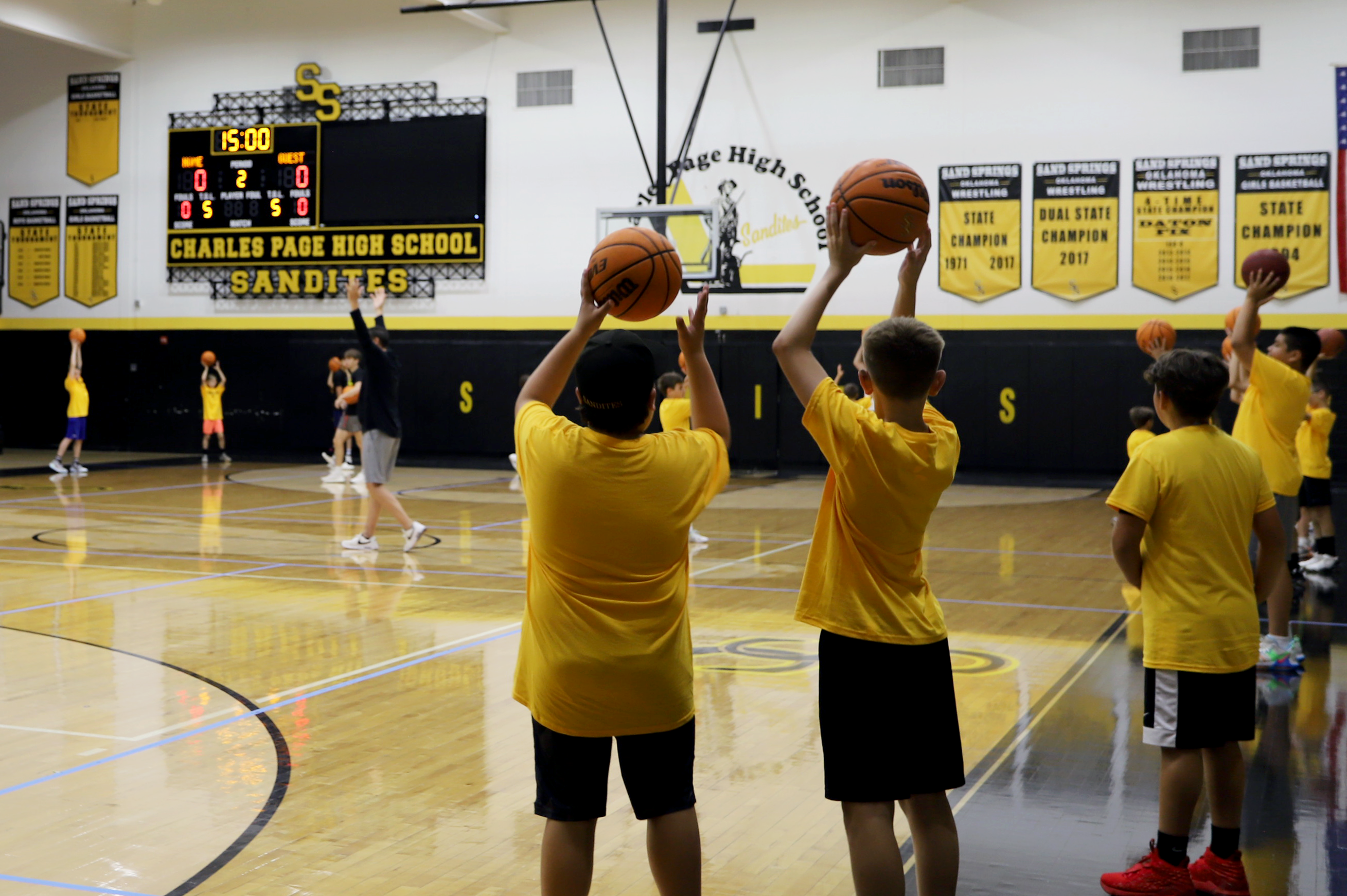 Elementary students in basketball gymnasium hold basketballs in the air at basketball camp