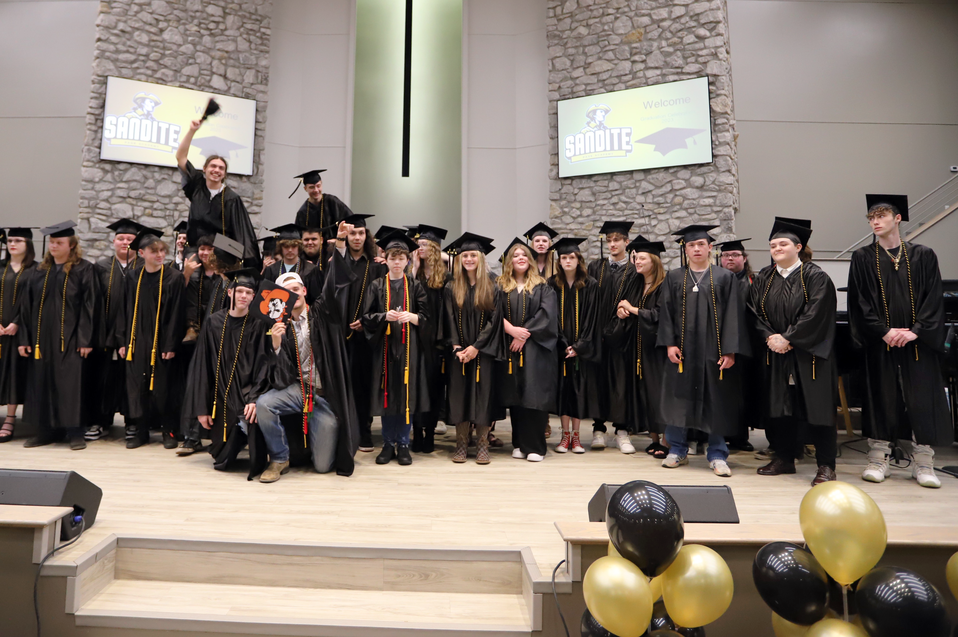 Students in black graduation caps and gowns smiling on stage