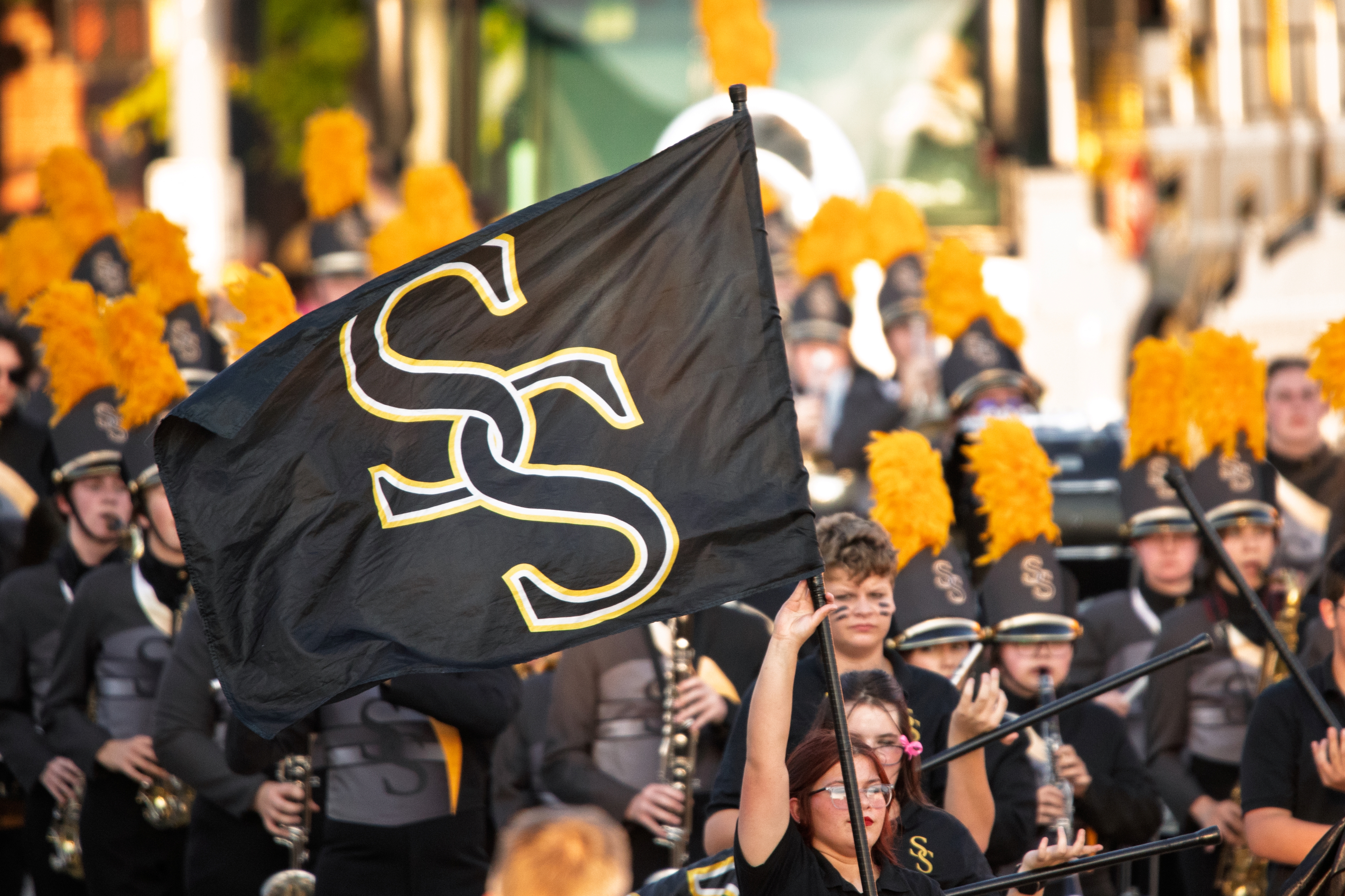 A marching band performs outdoors as a student waves a large black flag featuring interlocking gold “SS” letters. Band members in gray uniforms with bright yellow plumes on their hats play instruments in the background.