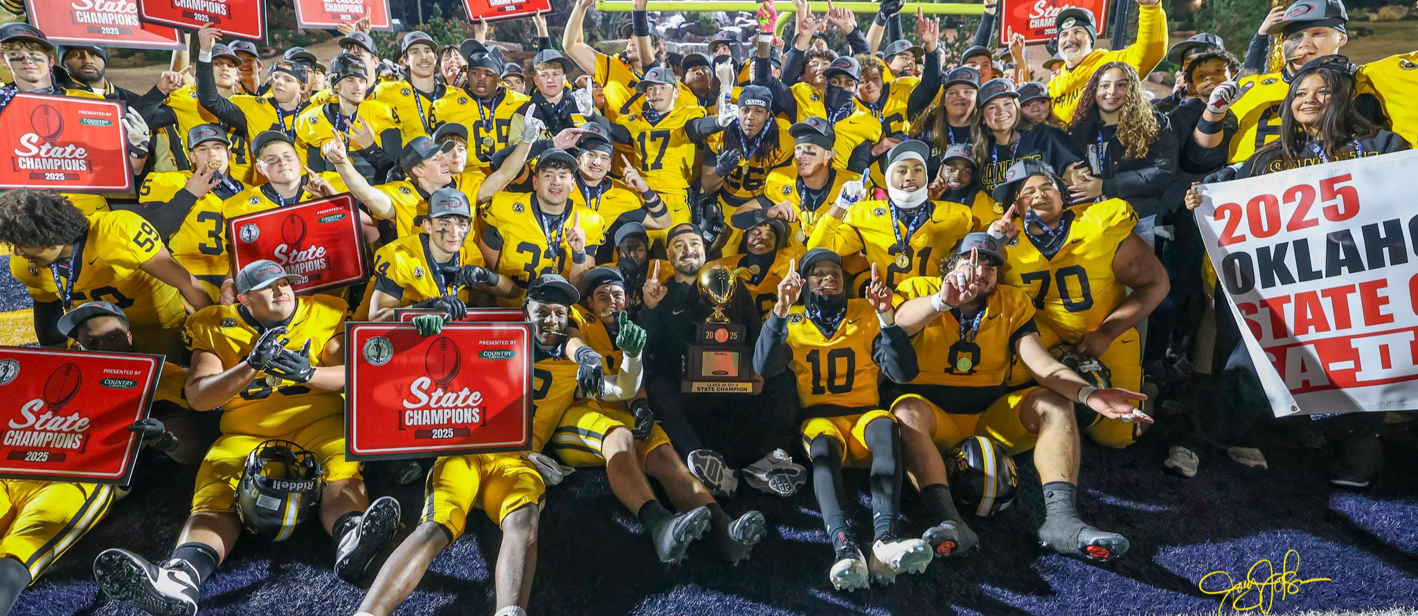 Sand Springs football team celebrates a 2025 state championship on the field, holding signs and a trophy.