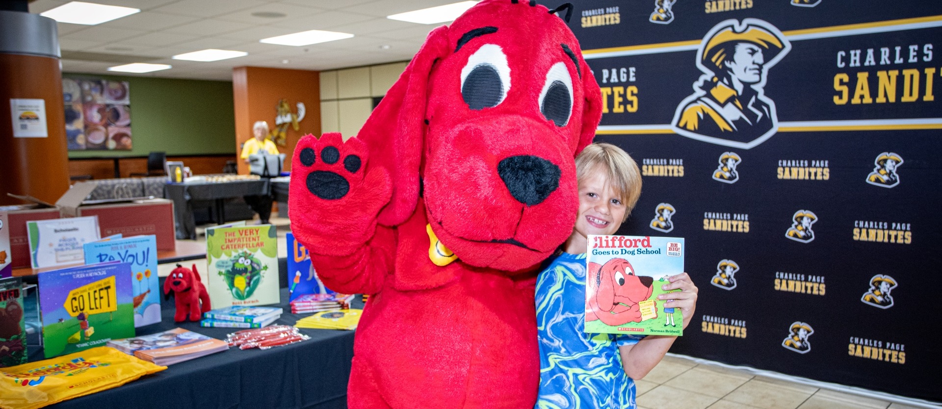 Student smiles while posing beside the Clifford the Big Red Dog mascot and holding a copy of “Clifford Goes to Dog School.” A display table filled with children’s books and a Charles Page Sandites backdrop frame the scene during a school literacy event celebrating reading and books.