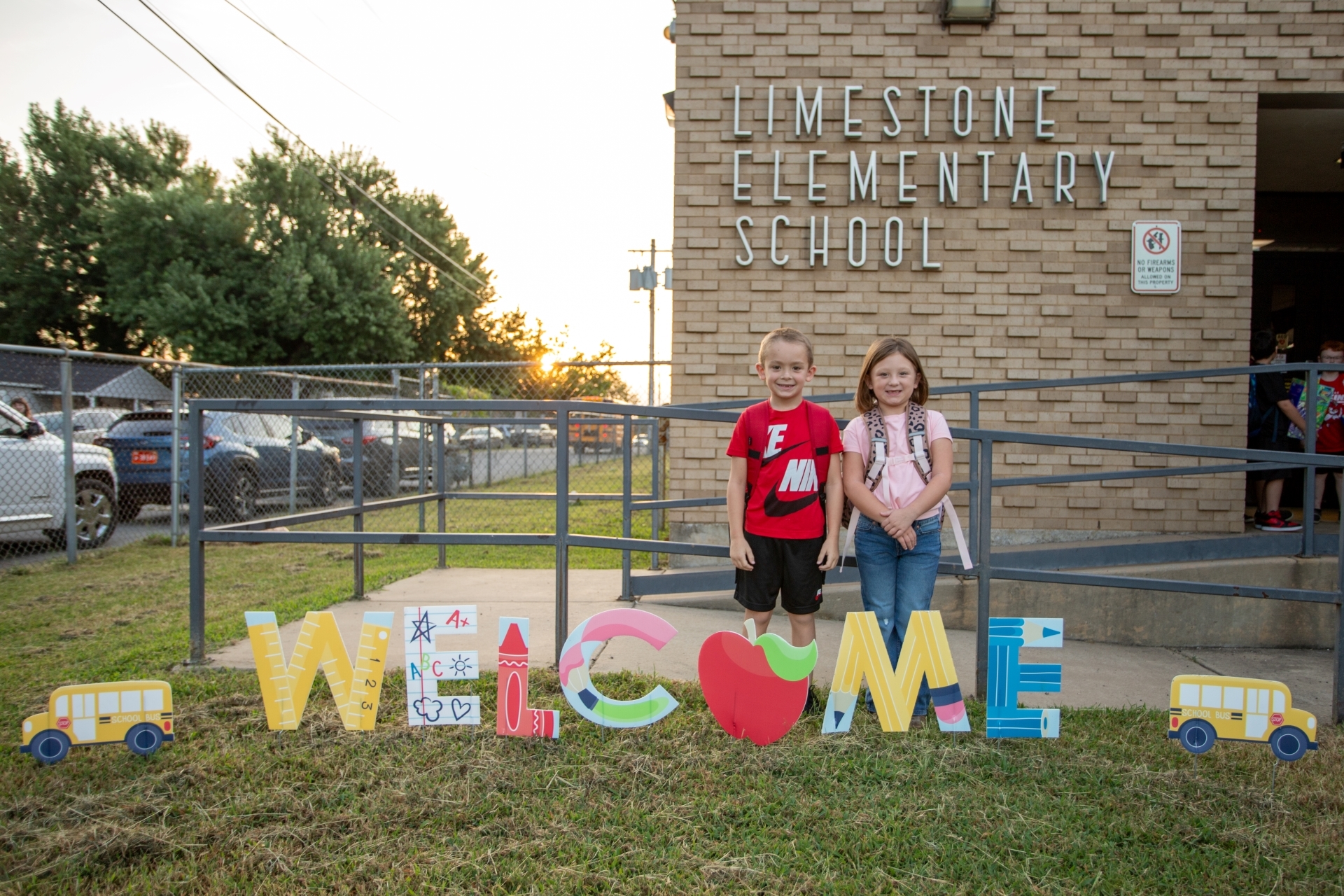 Two young students wearing backpacks pose behind a colorful "WELCOME" sign on the grass outside Limestone Elementary School.
