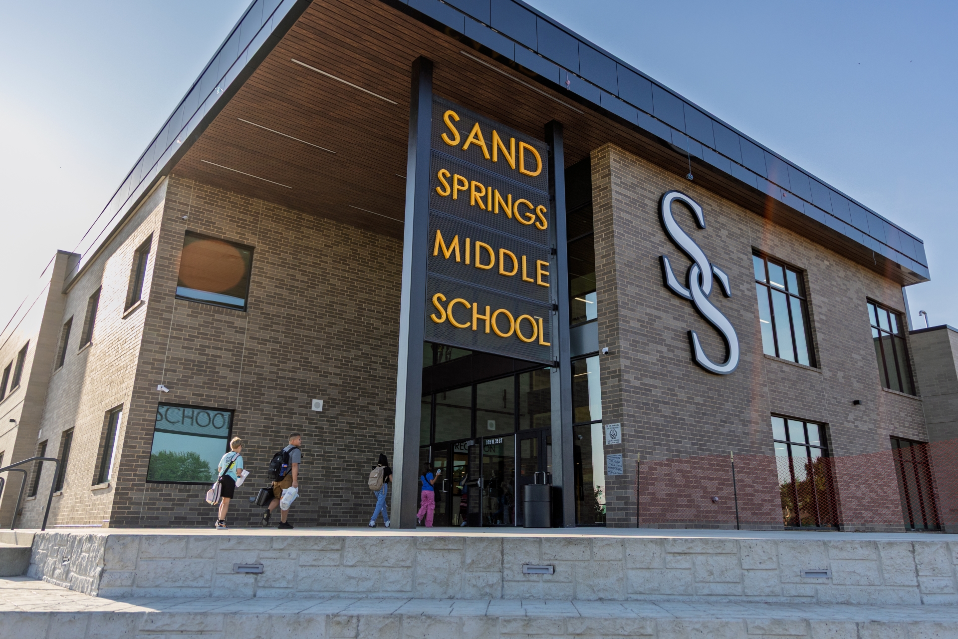 Students carrying backpacks walk toward the main entrance of Sand Springs Middle School, a modern brick building featuring vertical yellow letter signage and a large silver "SS" logo.