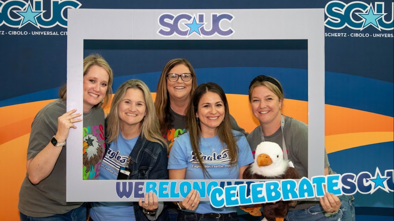 Five staff members smile behind an SCUC photo frame, holding a “We Belong. We Celebrate SCUC” sign with an eagle mascot in front of a district backdrop.