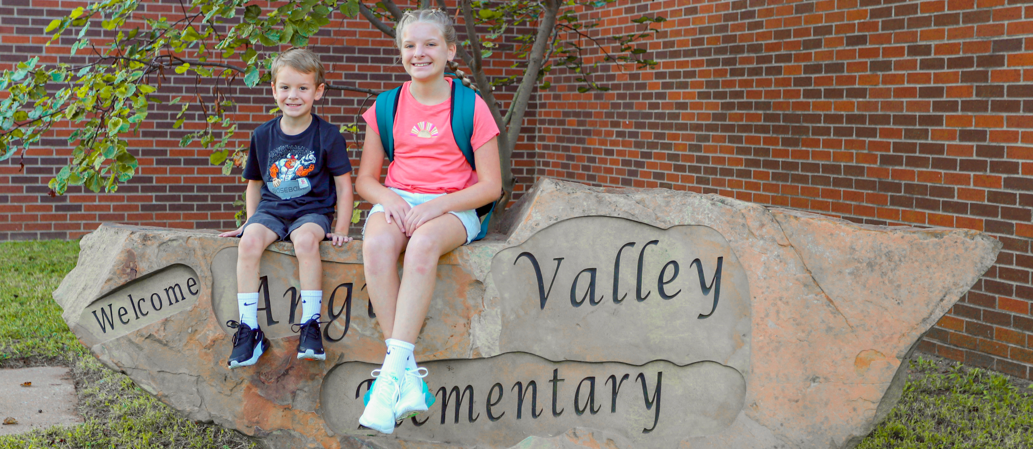 Two students sitting on rock with carved words: Welcome Angus Valley Elementary