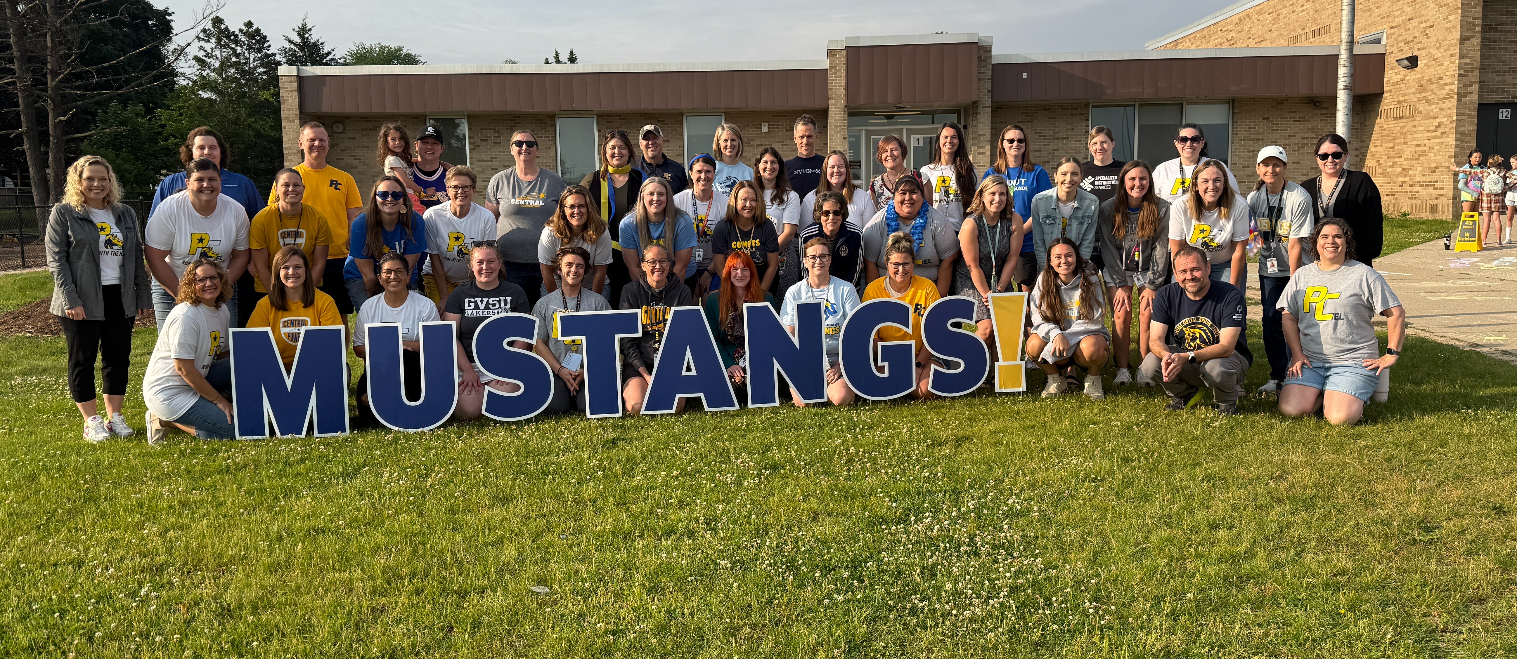 a large group of teachers pose behind Mustangs in front of a school