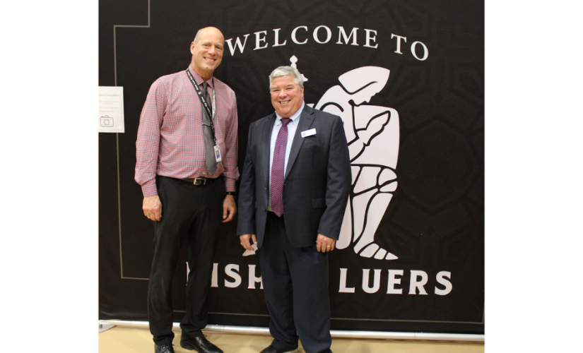 Principal Jim Huth in a suit standing to the right of a faculty member in front of the Bishop Luers logo and smiling
