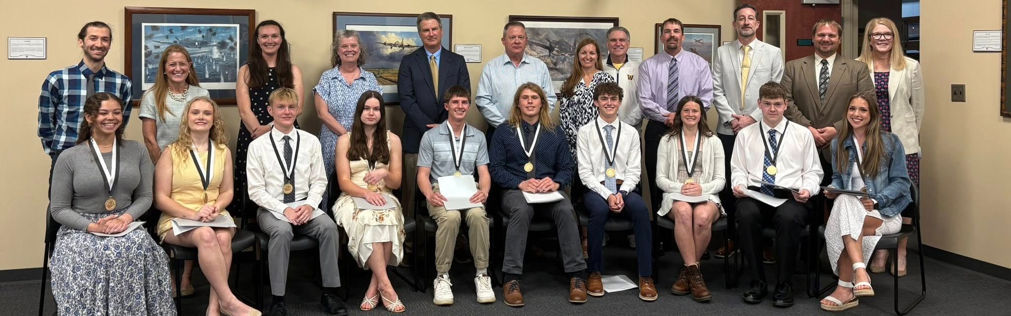 Ten students are seated in chairs in a row. Educators and administrators stand behind them. These are the top 10 high school seniors for the class of 2026.