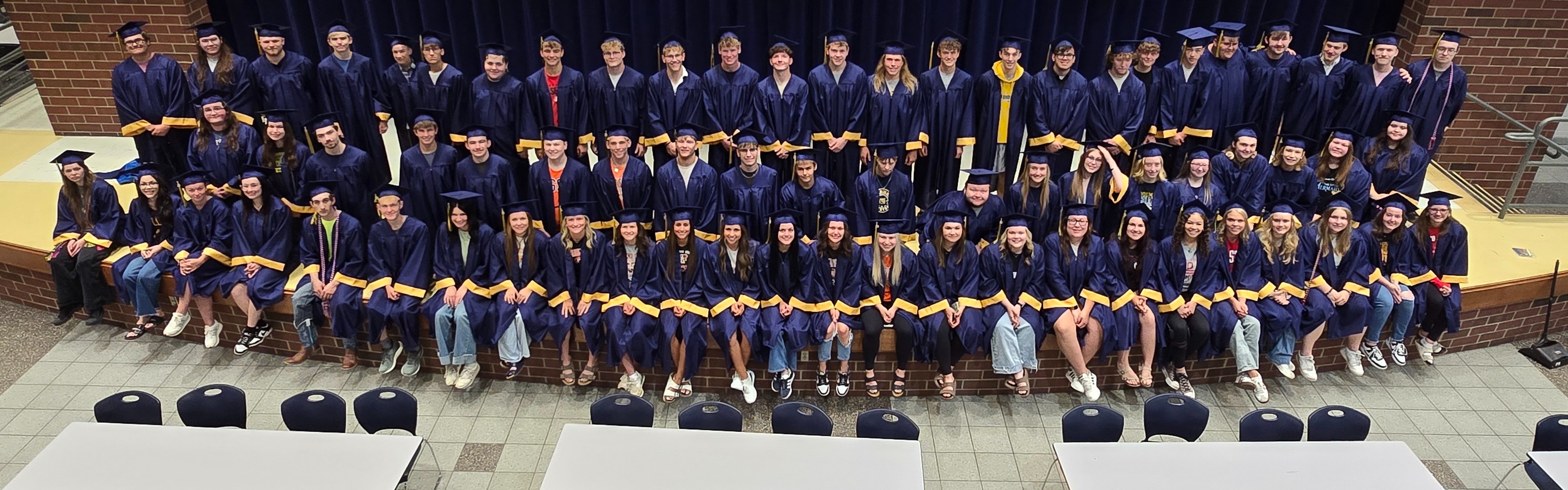 The Senior Class is lined up on the stage in their academic regalia (dark blue with gold trim on the sleeves) at the PK-8 building, looking up at the catwalk and posing for a picture
