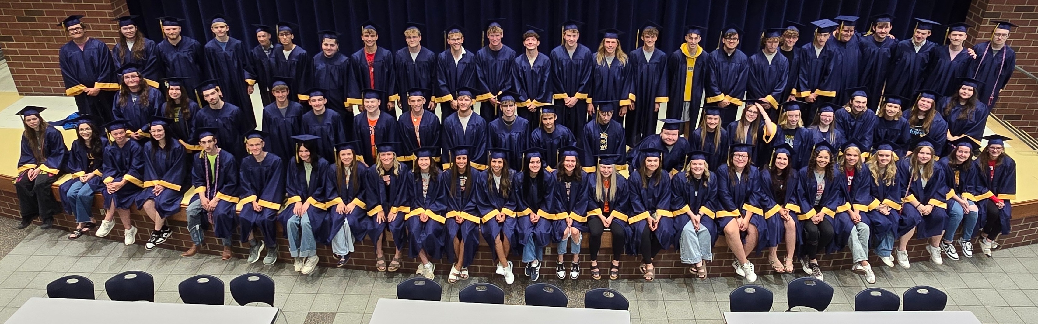 The Senior Class is lined up on the stage in their academic regalia (dark blue with gold trim on the sleeves) at the PK-8 building, looking up at the catwalk and posing for a picture
