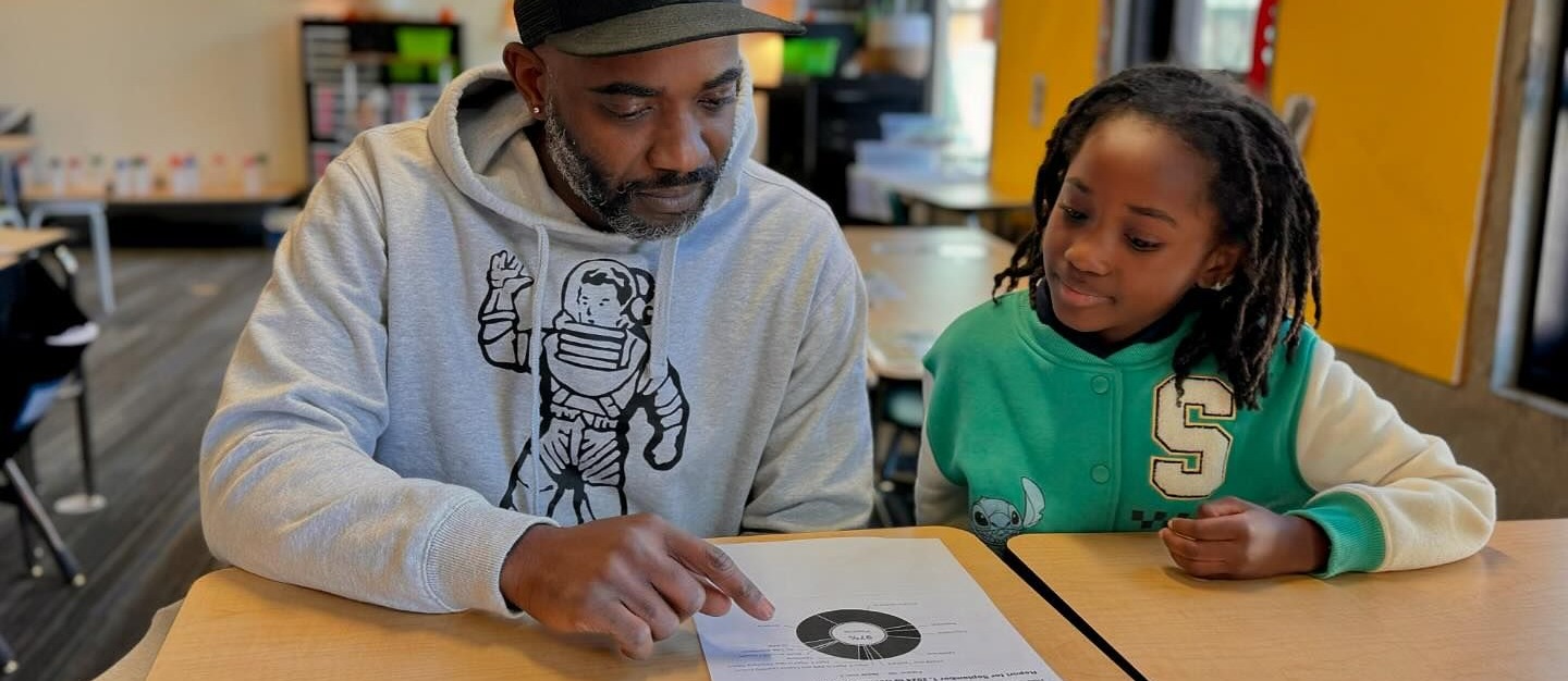 Family sits together reading learning assignments at a classroom desk