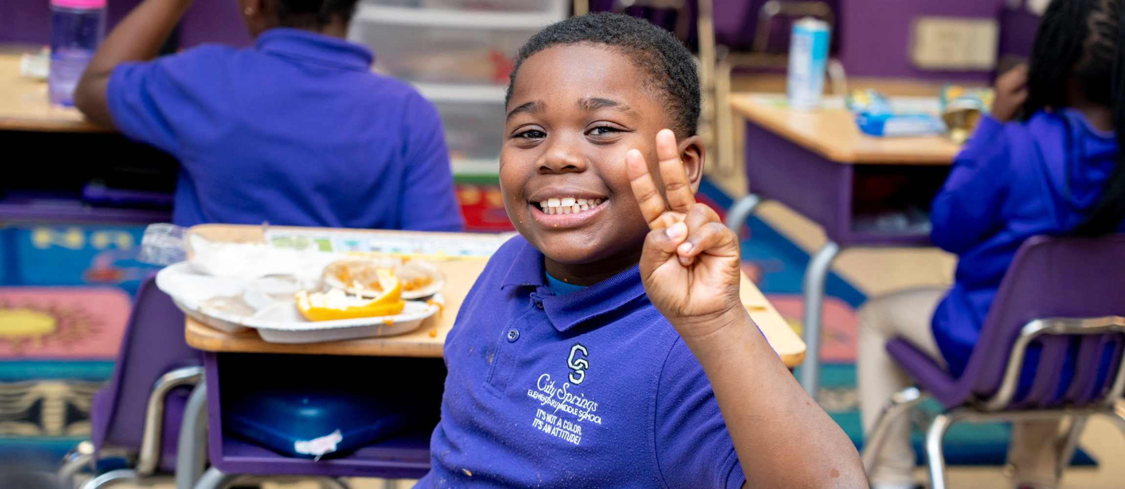 Smiling boy in a purple school uniform showing a peace sign while sitting at a classroom desk with a meal tray.
