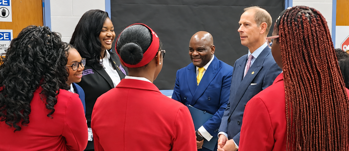 A group of professionally dressed students engaged in a lively conversation with The Duke of Edinburgh and City Schools CEO Dr. Dawson in an indoor setting with notice signs on the wall.