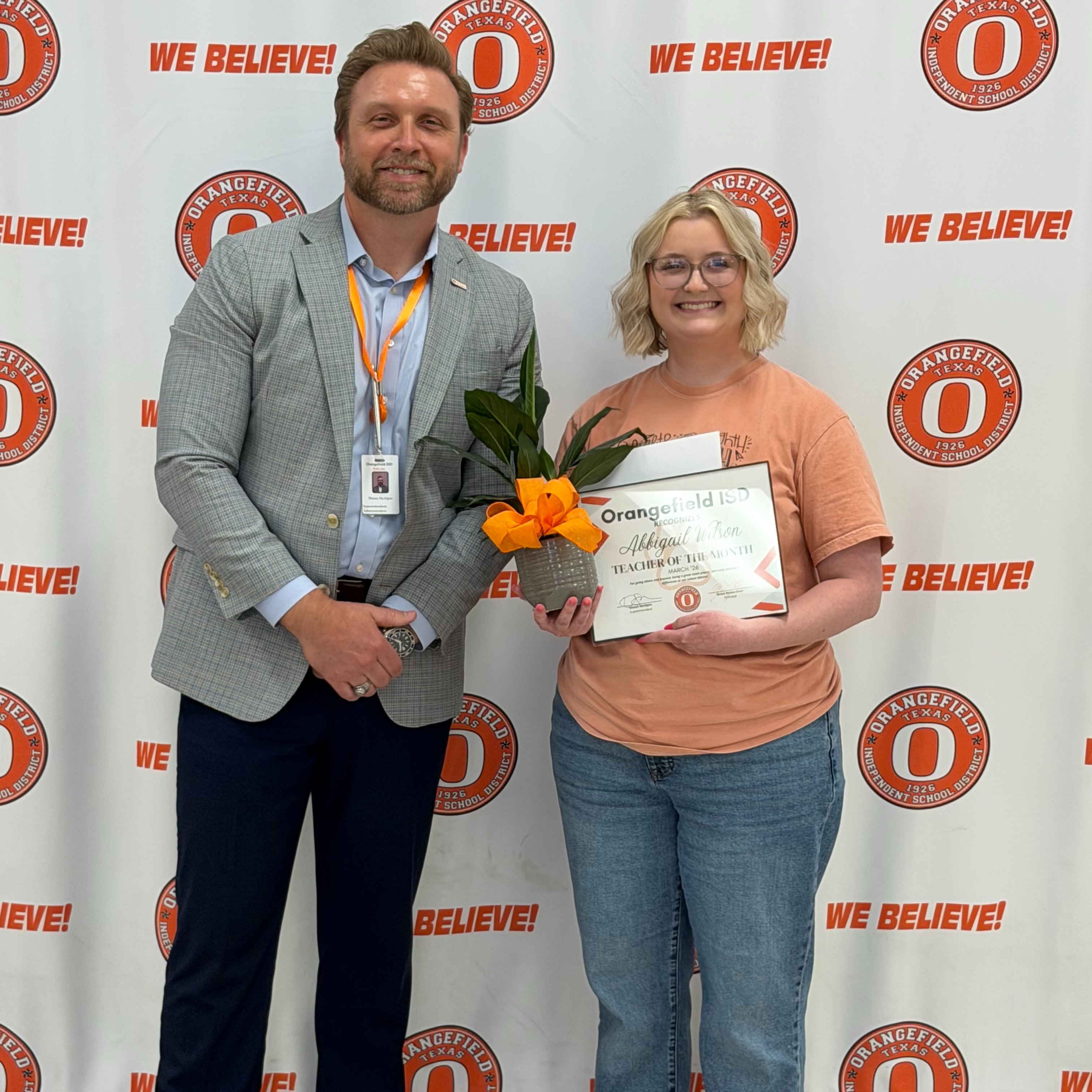 A man and woman stand before a white banner with orange logos and text reading "We Believe!". The woman holds a certificate, smiling.