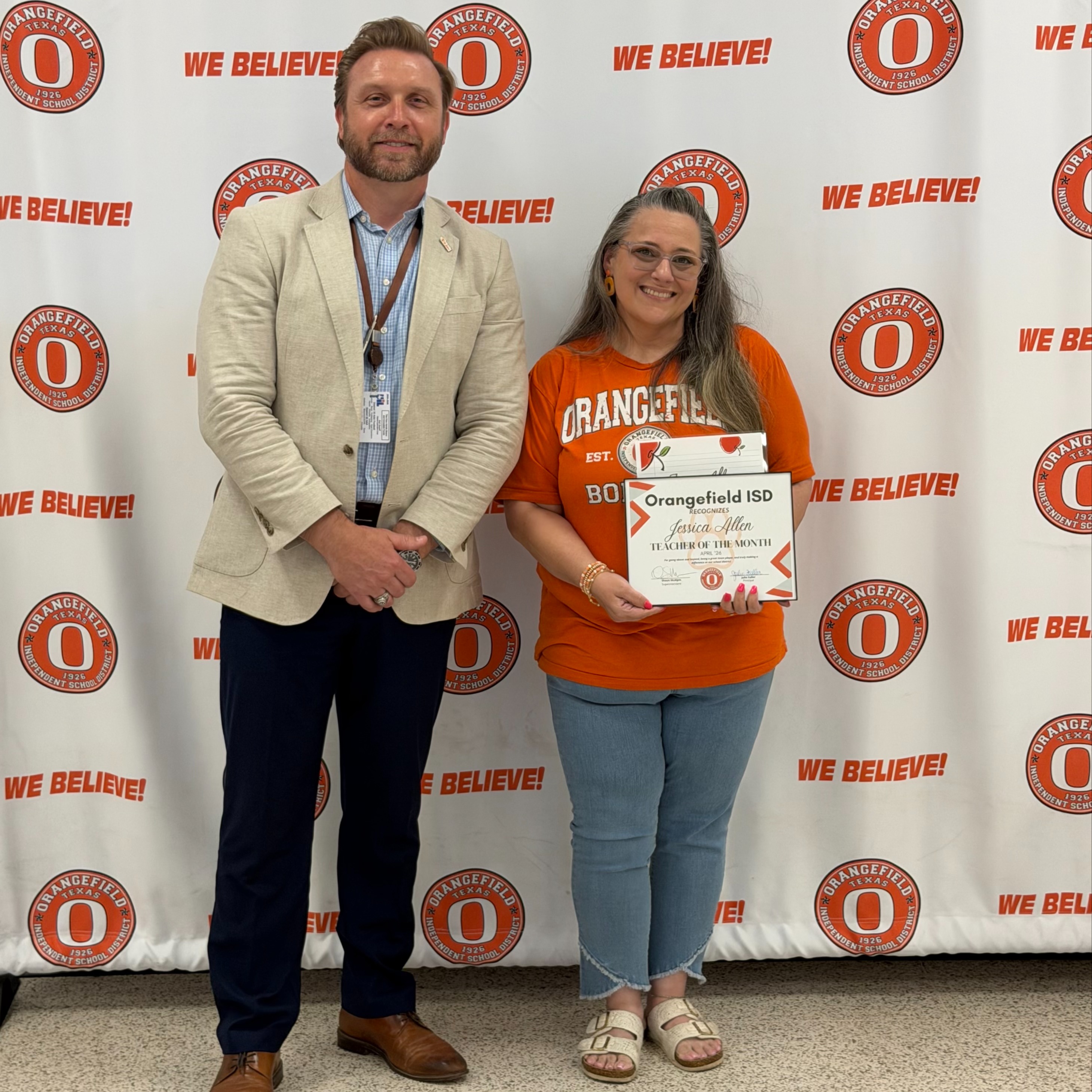 A man and a woman stand smiling before a white backdrop with orange circles and the text "WE BELIEVE!" The woman holds a plaque.