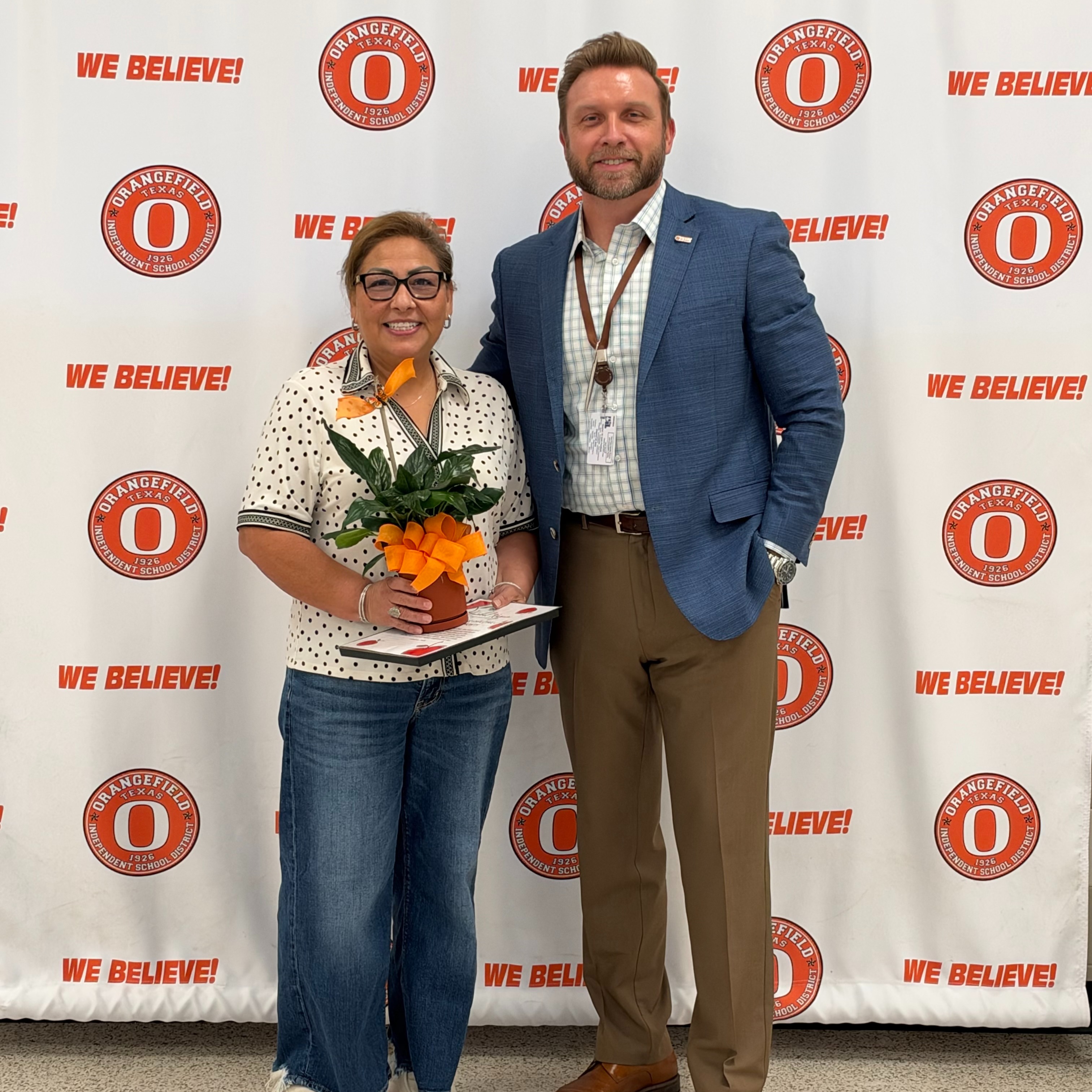 Two adults stand in front of a wall with red, white, and orange branding. A woman holds a potted plant and a book. A man wears a watch.