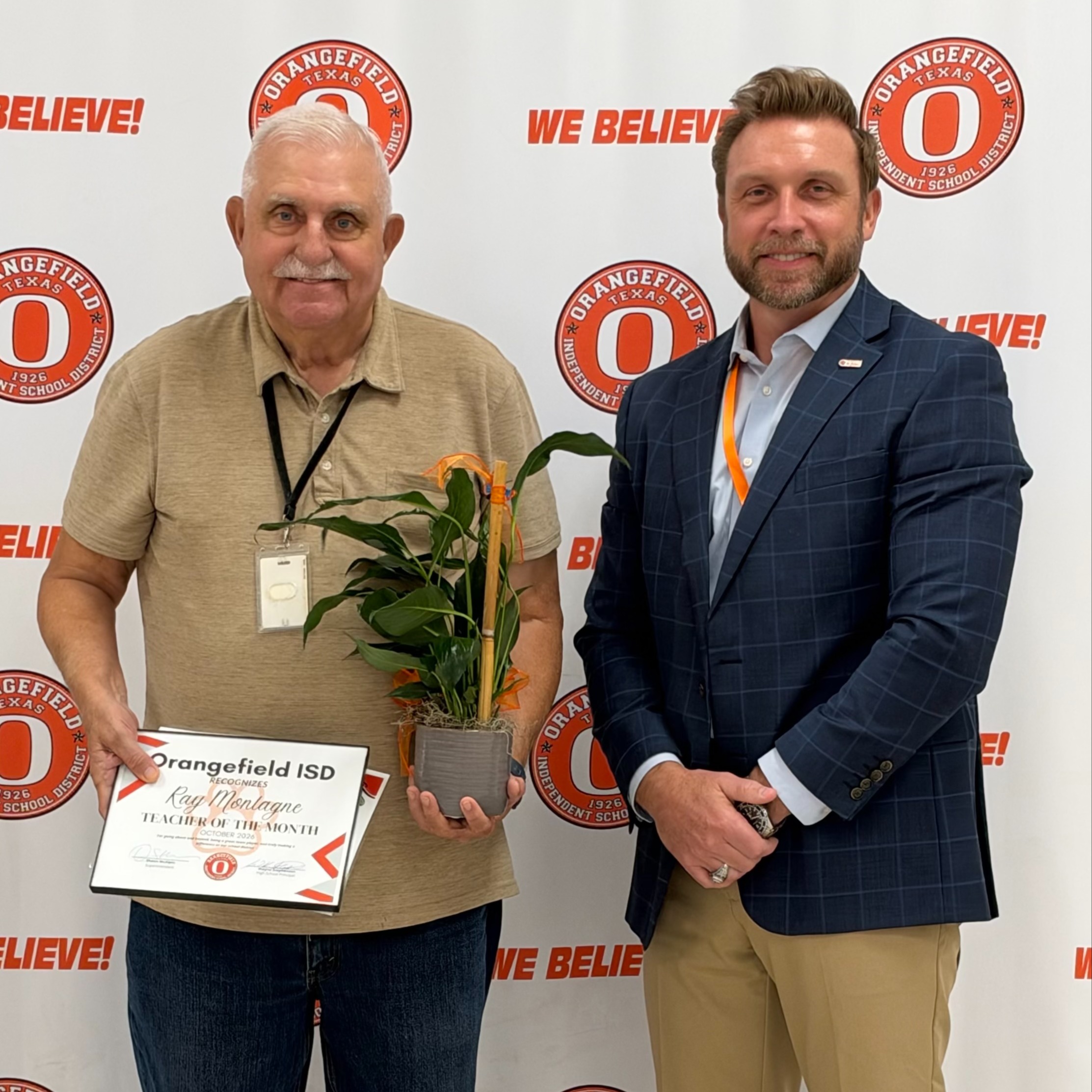 Two men in formal attire standing in front of a backdrop with the text "We Believe!" One man holds a certificate and a potted plant.