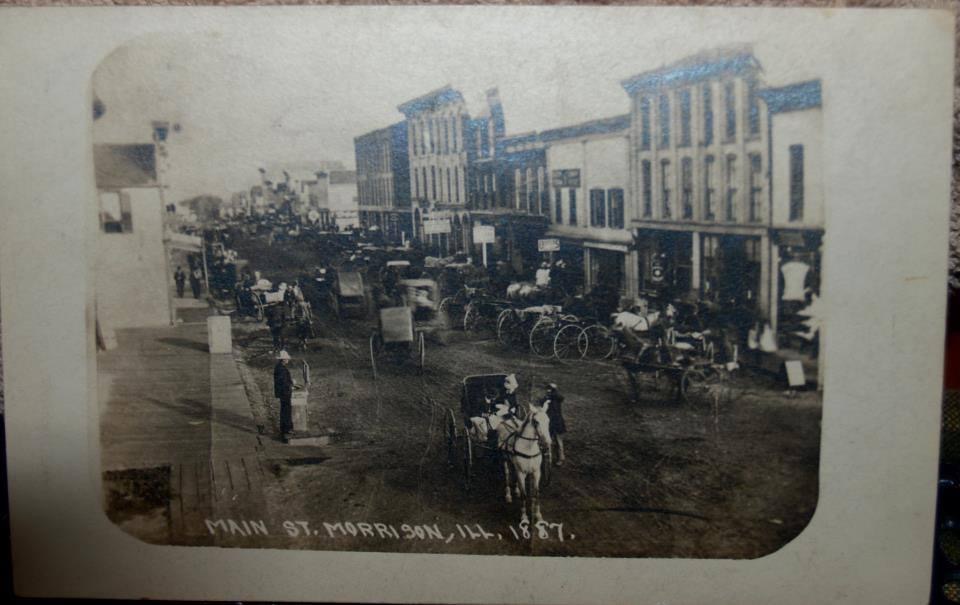 black and white photo of main street with horse drawn carriages parked outside of the buildings