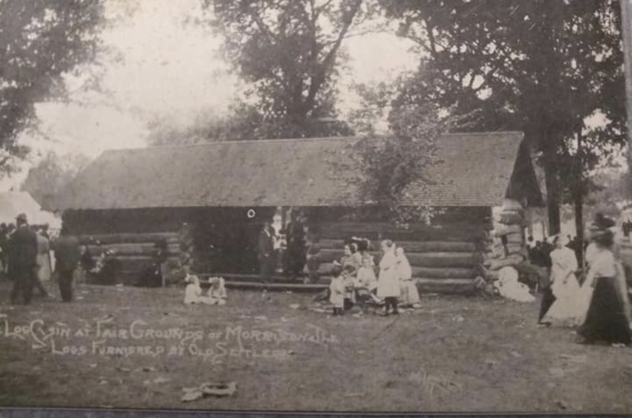 black and white photo of a log cabin with people gathered on the lawn out front