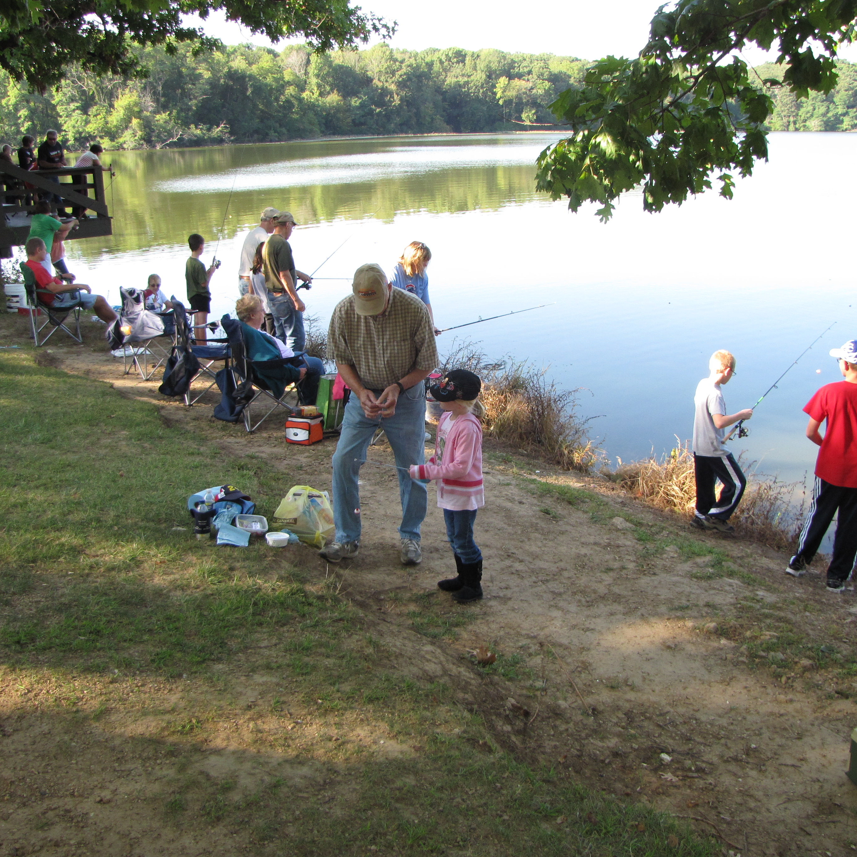 people line the bank of a lake holding fishing poles