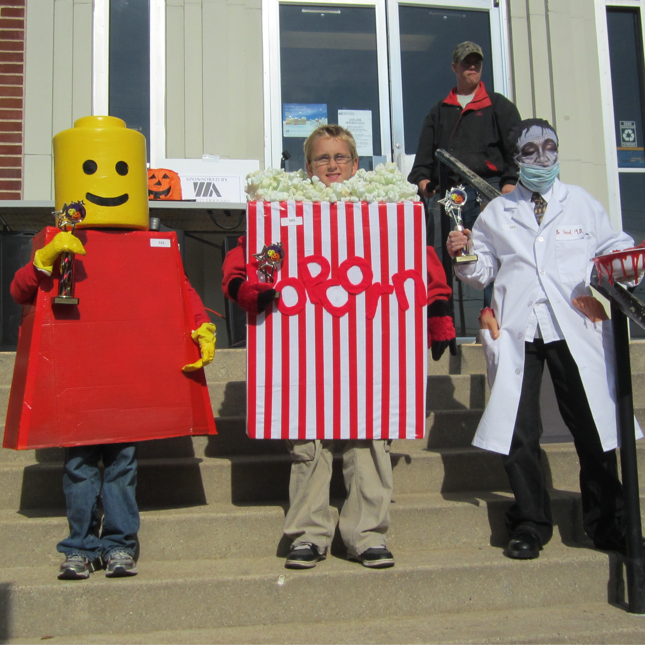 kids dressed as a lego popcorn and frankenstein stand with trophies on the steps of a building