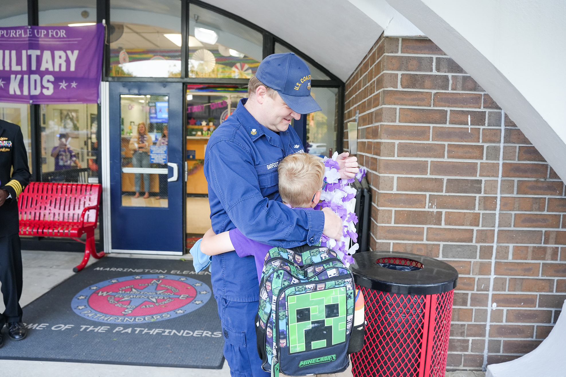 Man in uniform hugs a child with a backpack. Brick wall with a trash can and banner behind them.