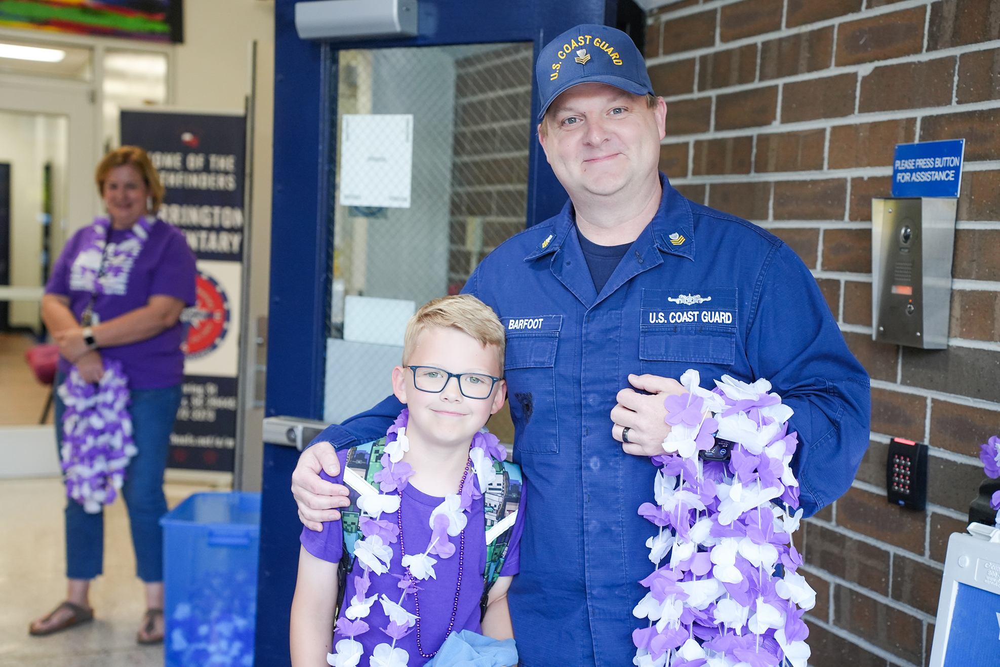 A man in navy blue uniform holds a lei around a child. The man wears a cap, glasses, and a watch. The child wears a lei and glasses.
