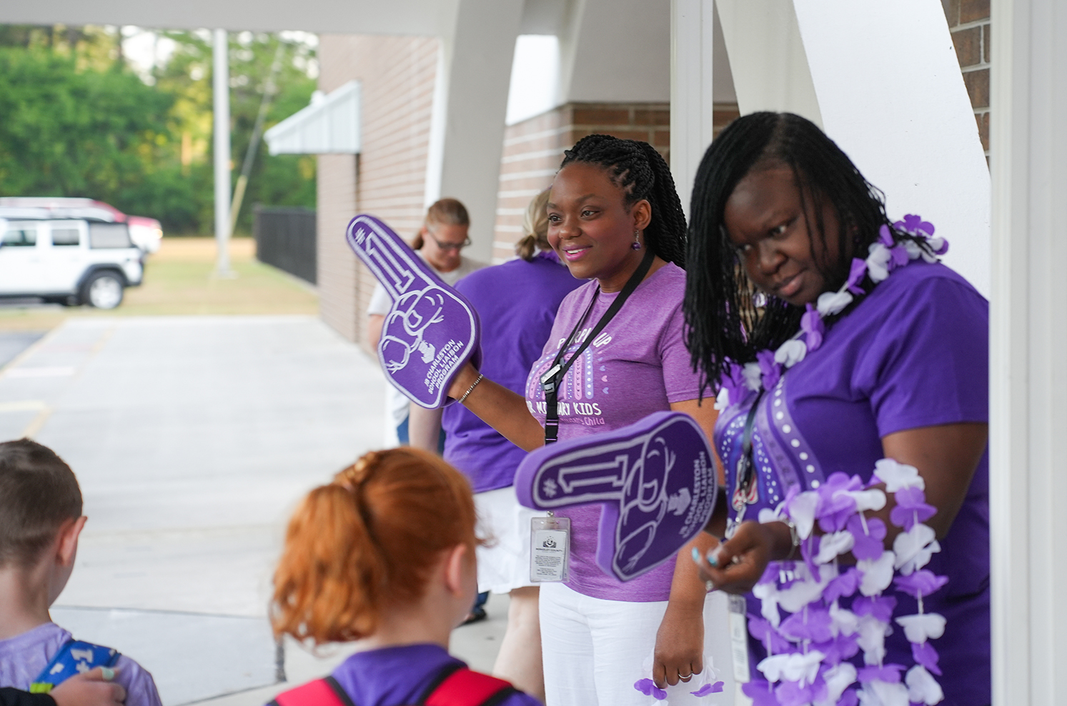 Several adults and children in matching purple shirts stand near a building. One adult holds a purple paw.