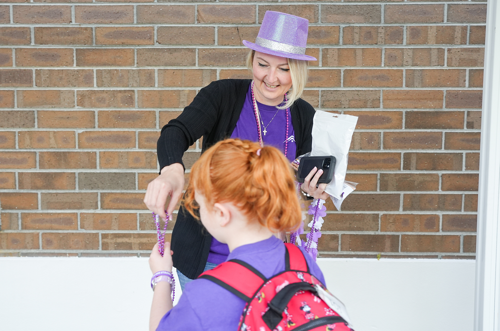 A smiling woman with a purple hat hands a purple ribbon to a child with red hair and a red backpack, against a brick wall.