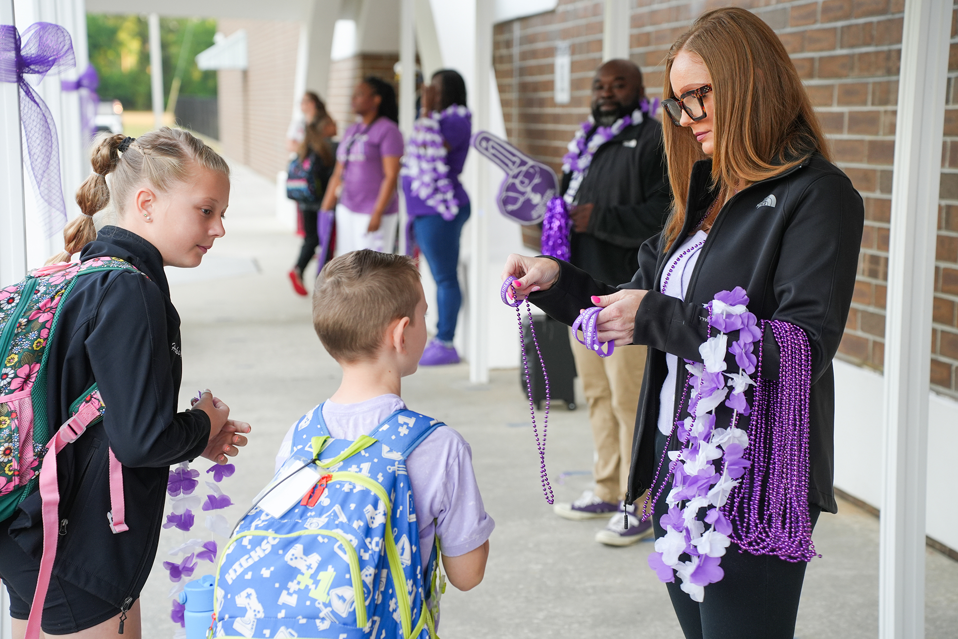 A woman hands a decorative ribbon to a child in a school corridor. Behind them, more children and adults stand.