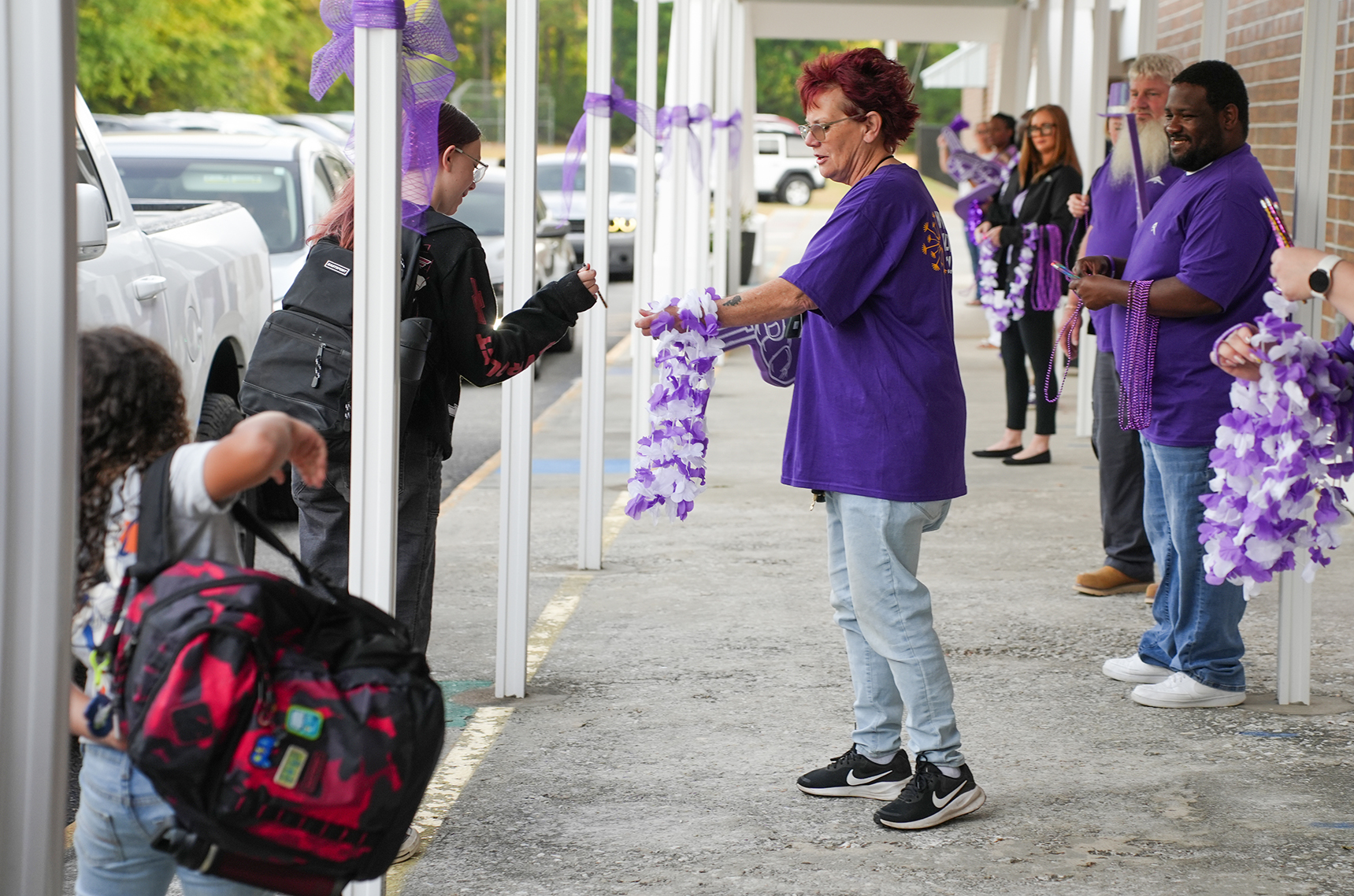 Purple-shirted adults stand on a sidewalk, distributing ribbons. A child holding a backpack stands nearby.