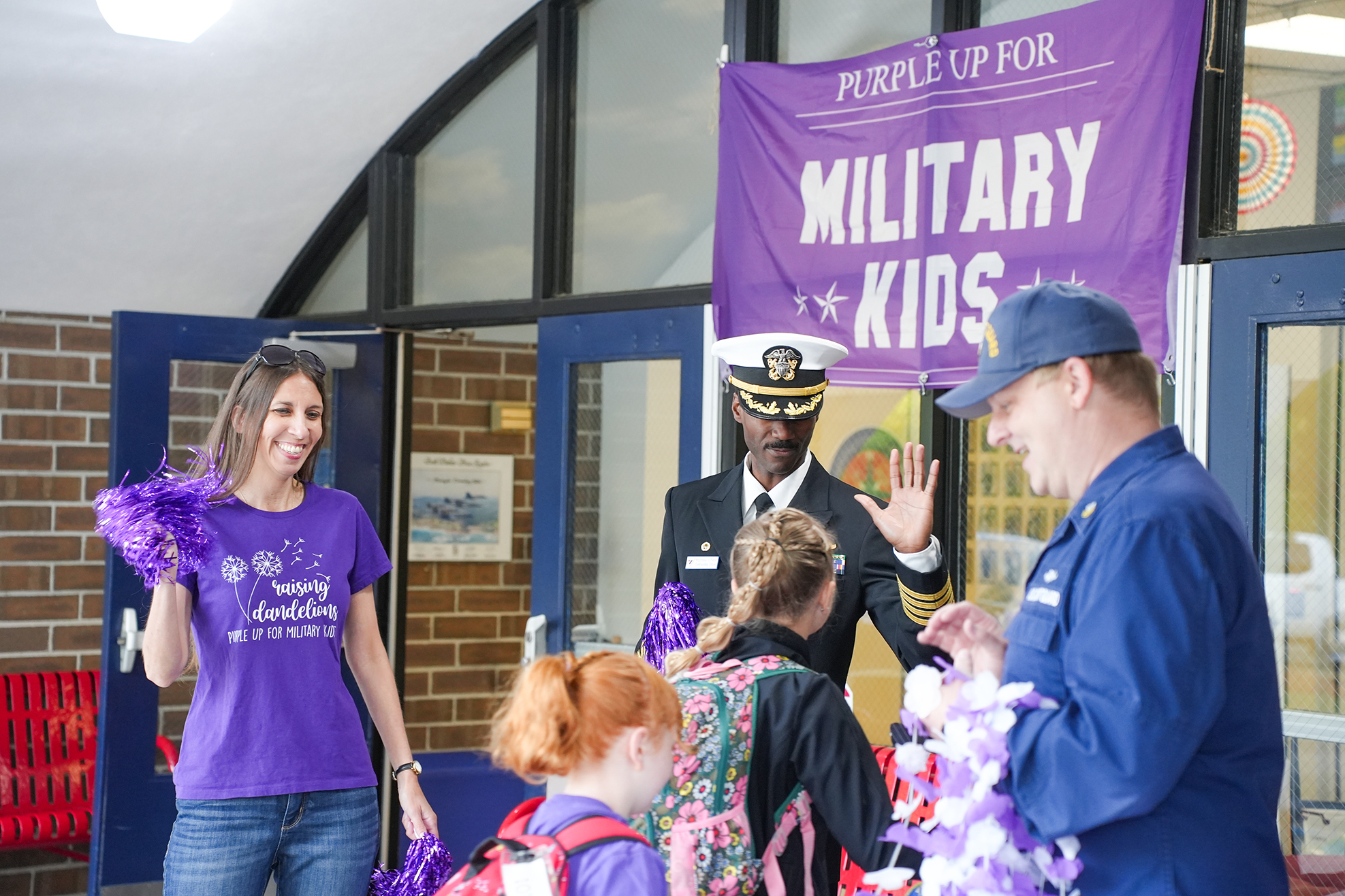Military personnel in uniform outside a school building. A banner reads "Purple Up for Military Kids."